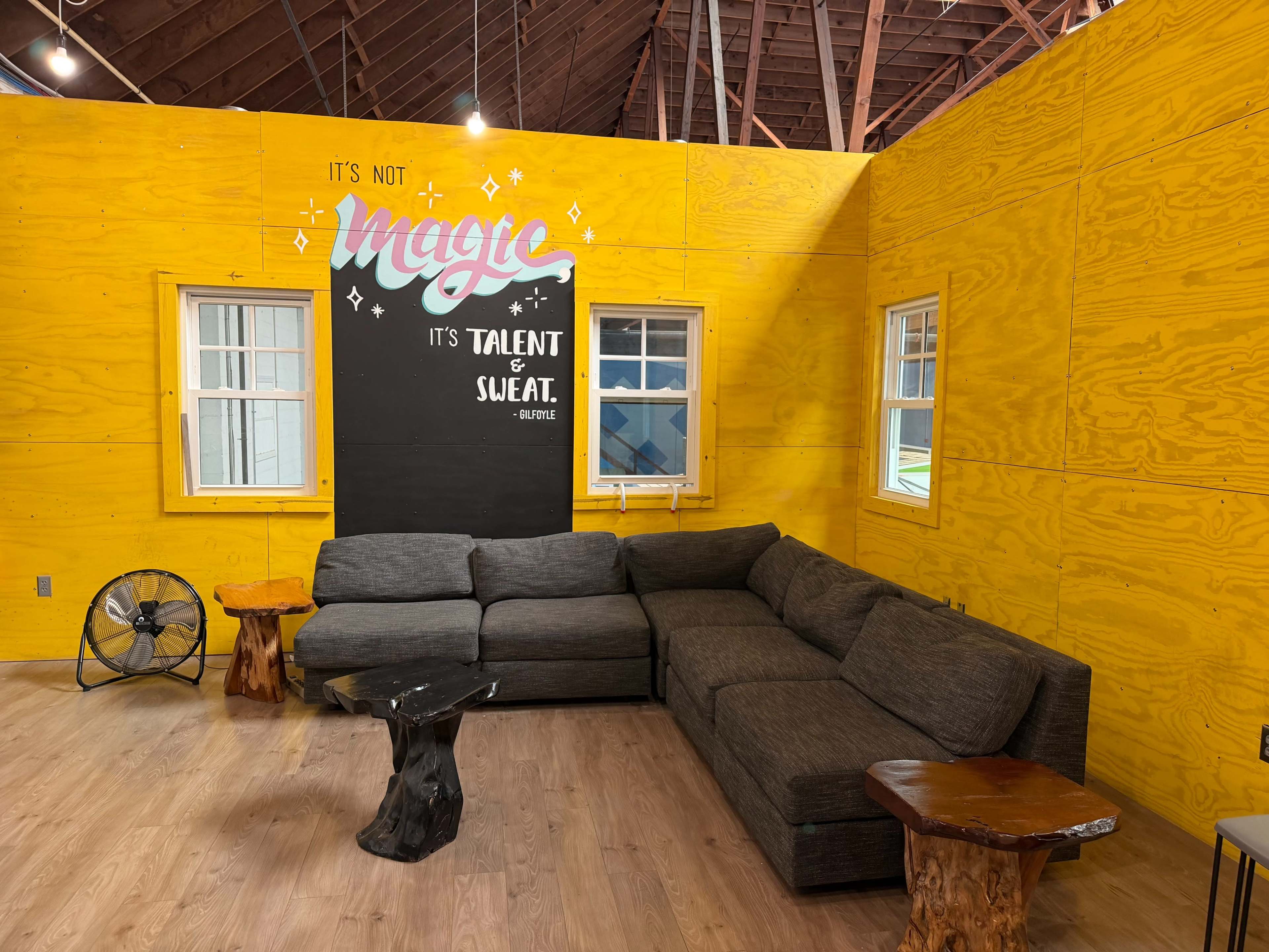 The image shows a cozy lounge area featuring a dark gray sectional sofa, wooden side tables, and a yellow wall with a motivational mural.
