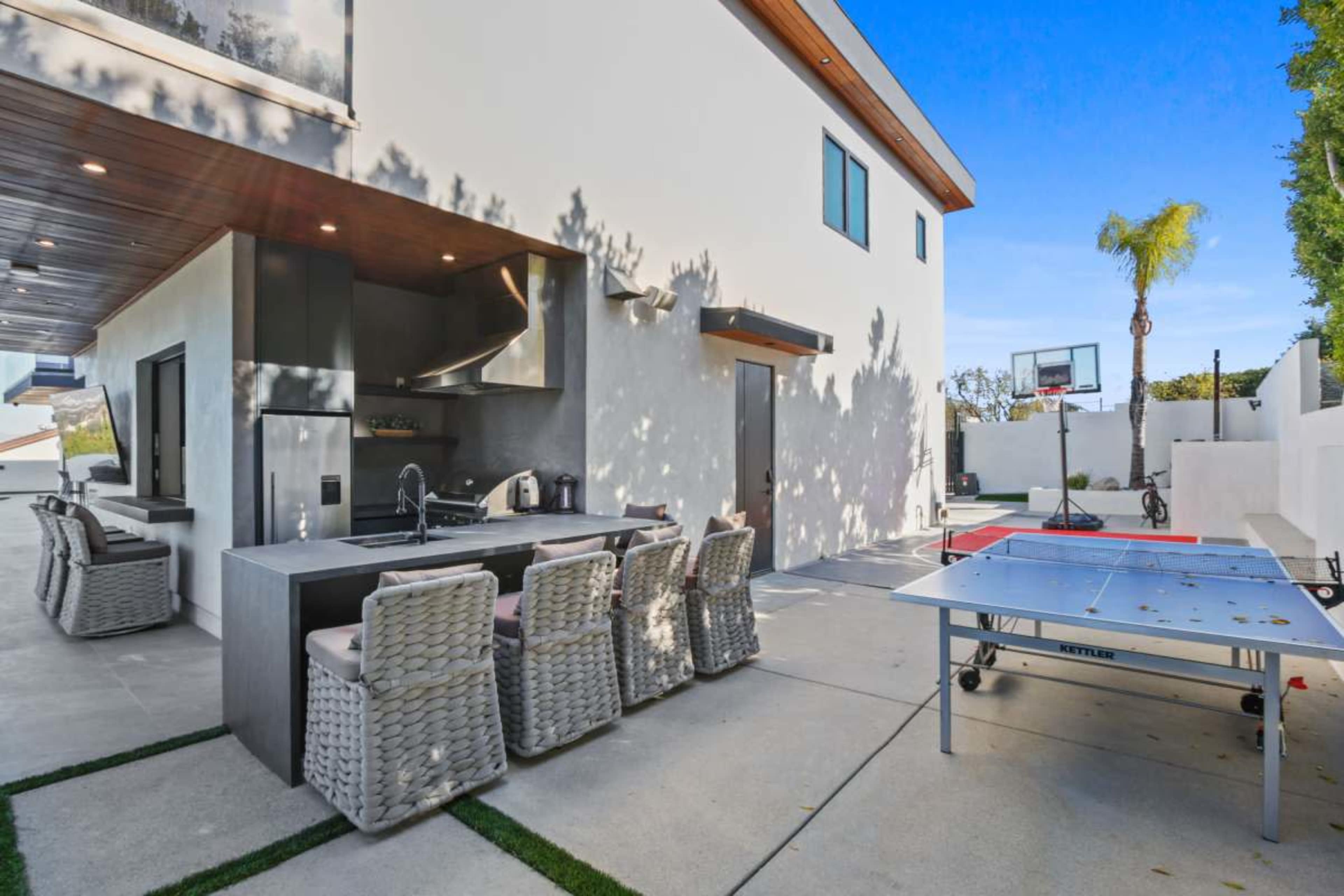The image shows a modern outdoor space featuring a large patio with a kitchen area, seating, and a ping pong table beside a basketball hoop.