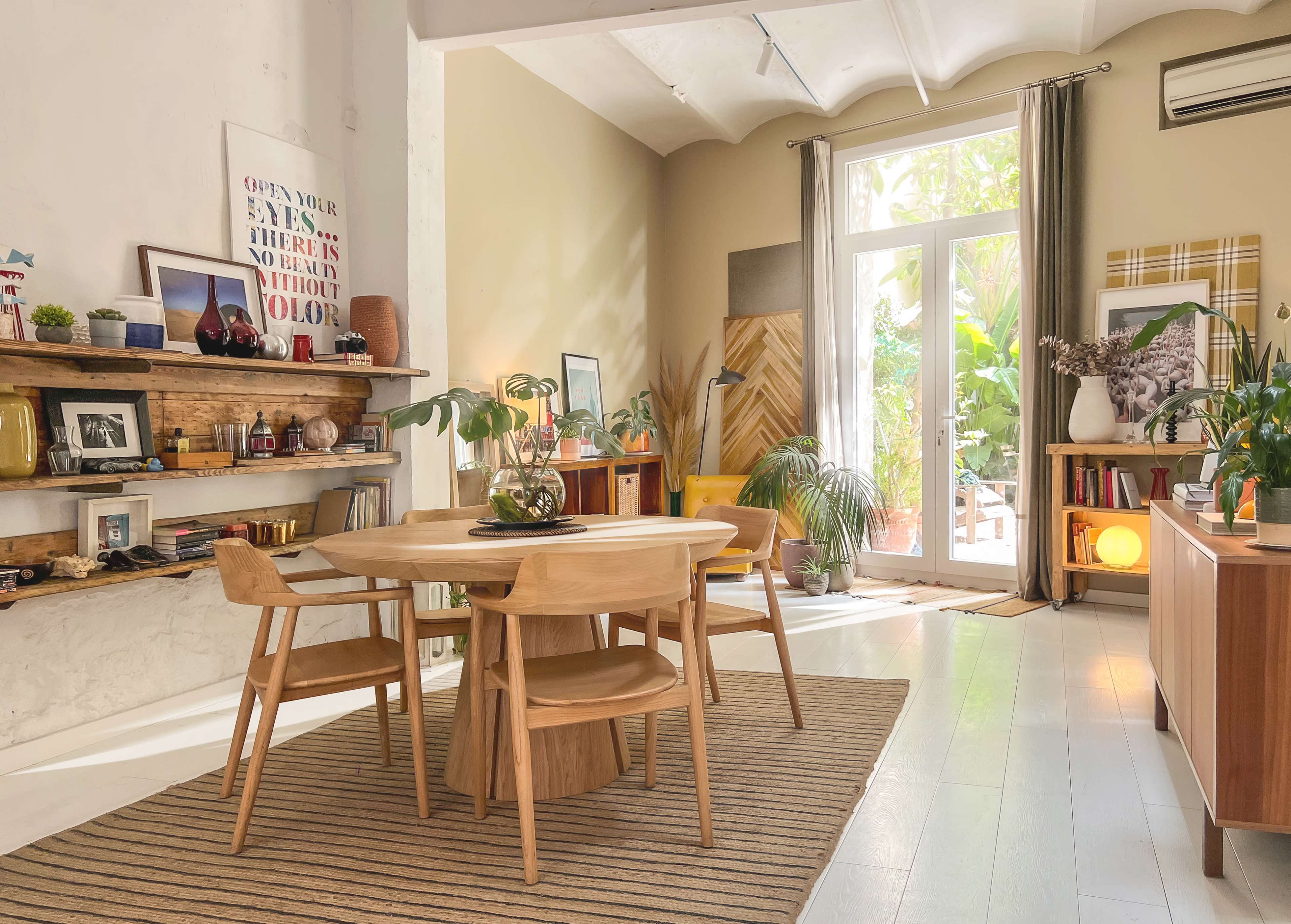 A wooden dining table surrounded by chairs is set in a well-lit room with a plant and shelves displaying various decorations against a backdrop of large windows.