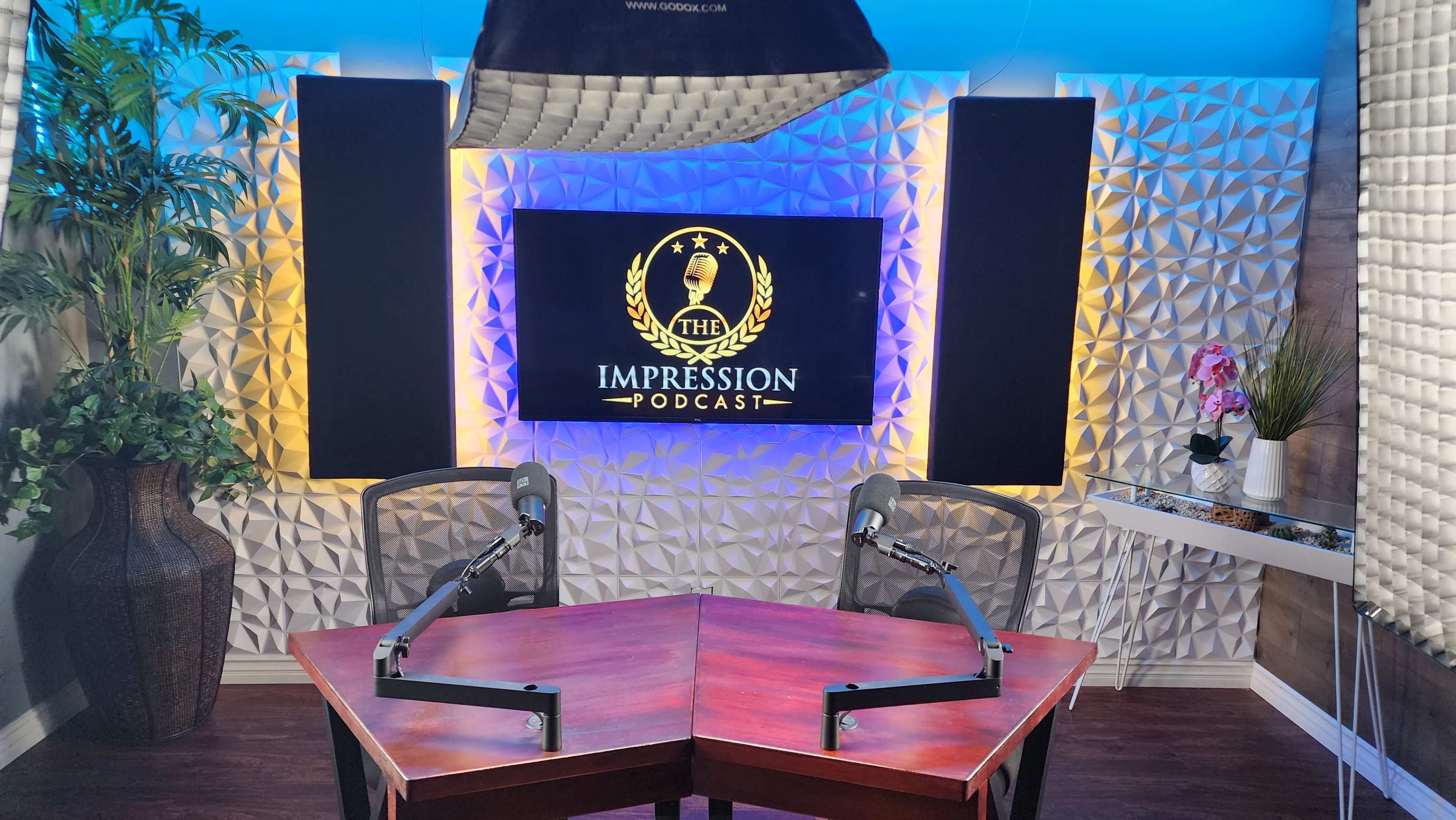 A podcast studio with a decorative wall, two mounted microphones on a wooden table, and a screen displaying "The Impression Podcast" in the background.
