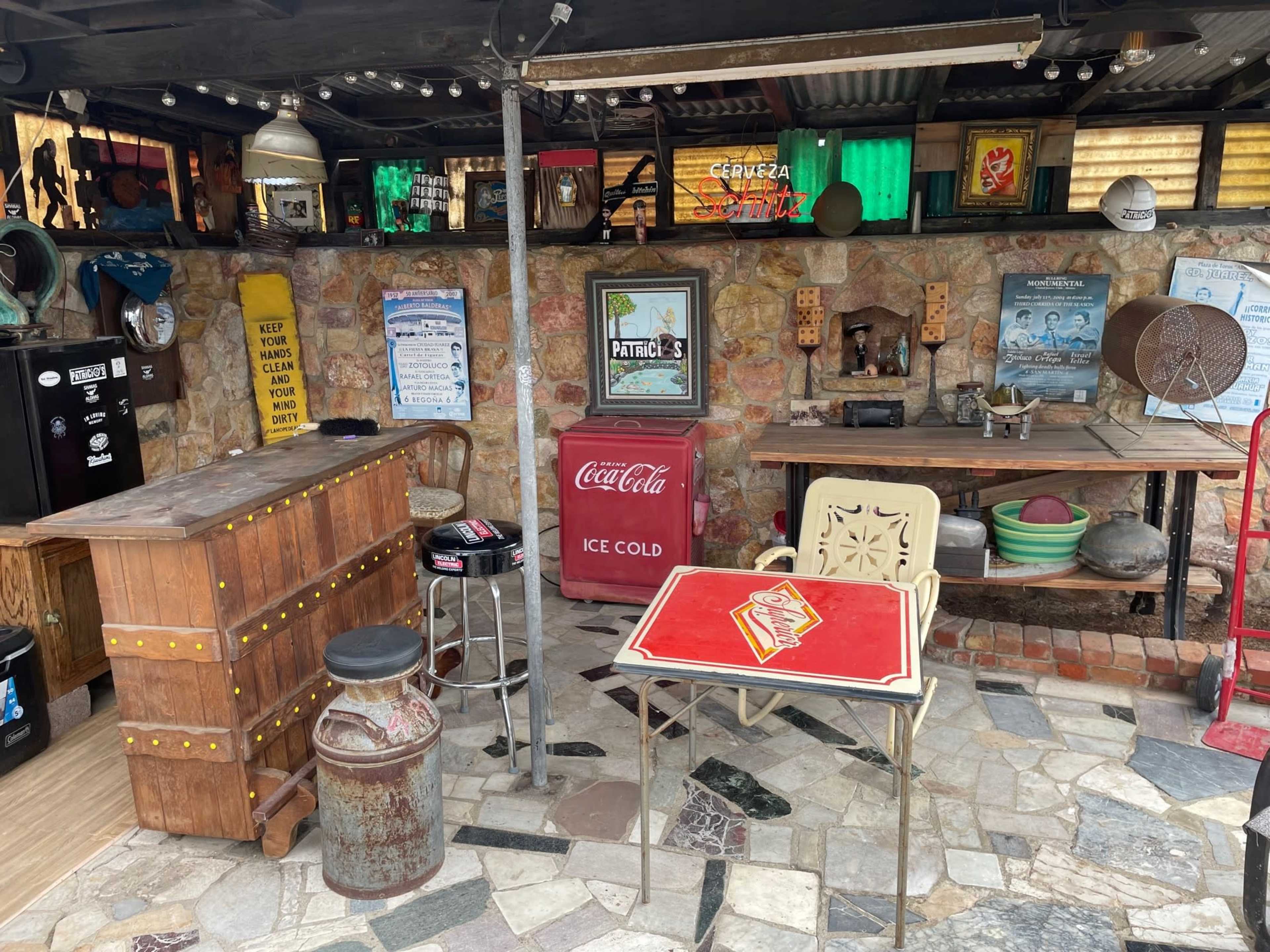 The image shows a rustic outdoor bar area featuring a wooden bar counter, assorted vintage decorations, a Coca-Cola ice chest, and various seating arrangements on a stone floor.