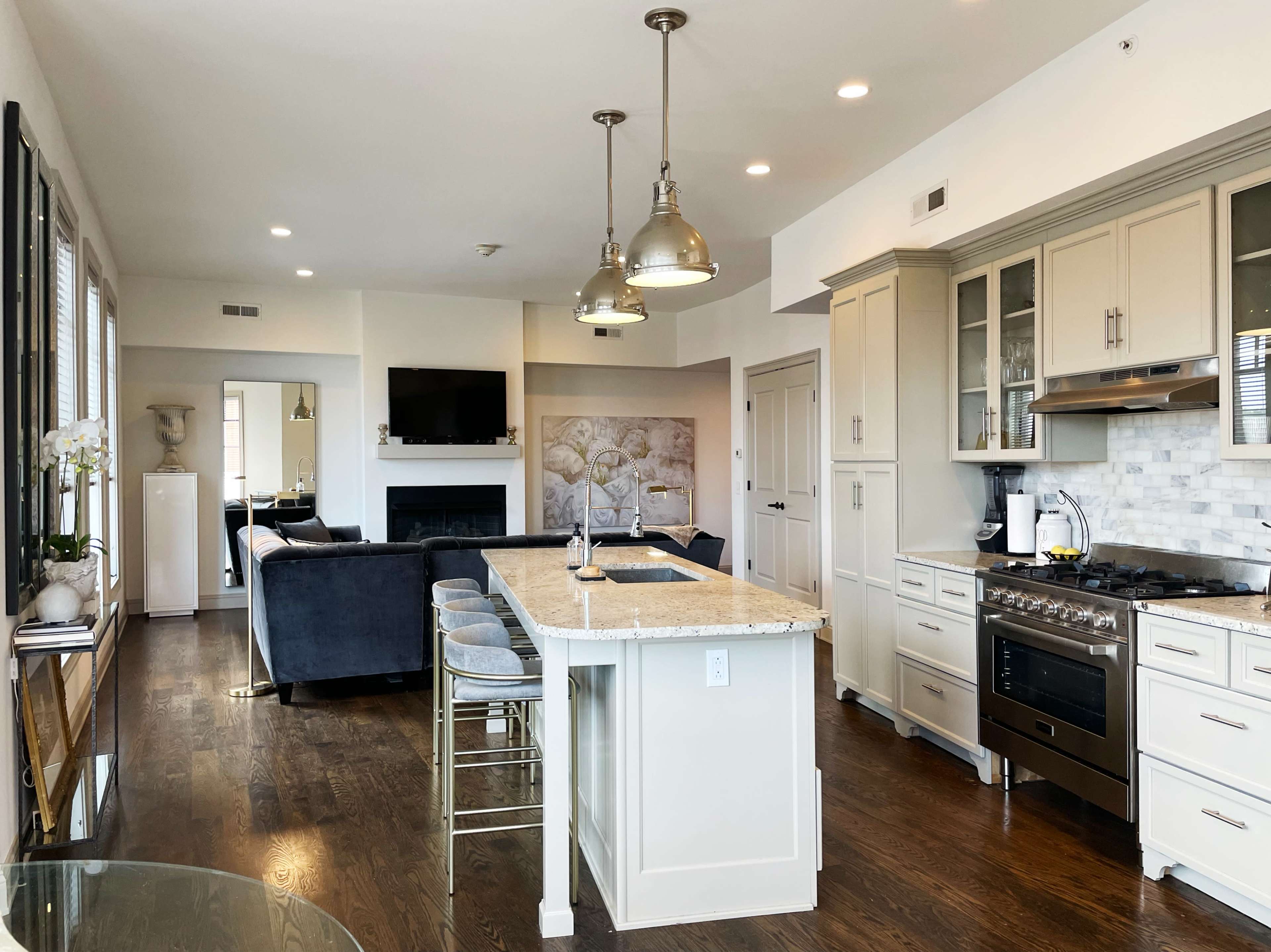 A modern kitchen with an island, featuring stainless steel appliances, a stone countertop, and a living area with a television in the background.