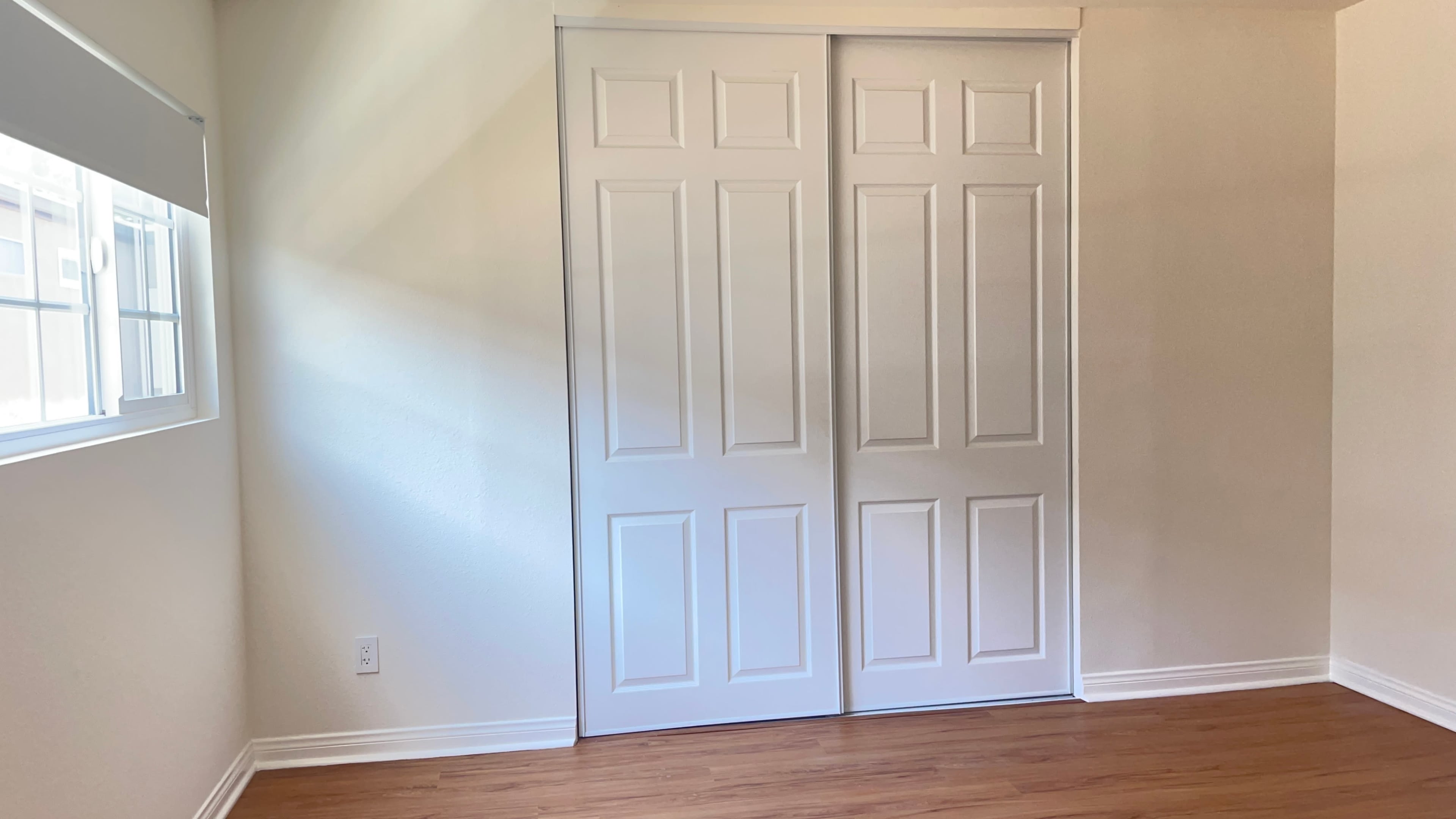 A bright room with light-colored walls, a wooden floor, and a double door closet on one side.