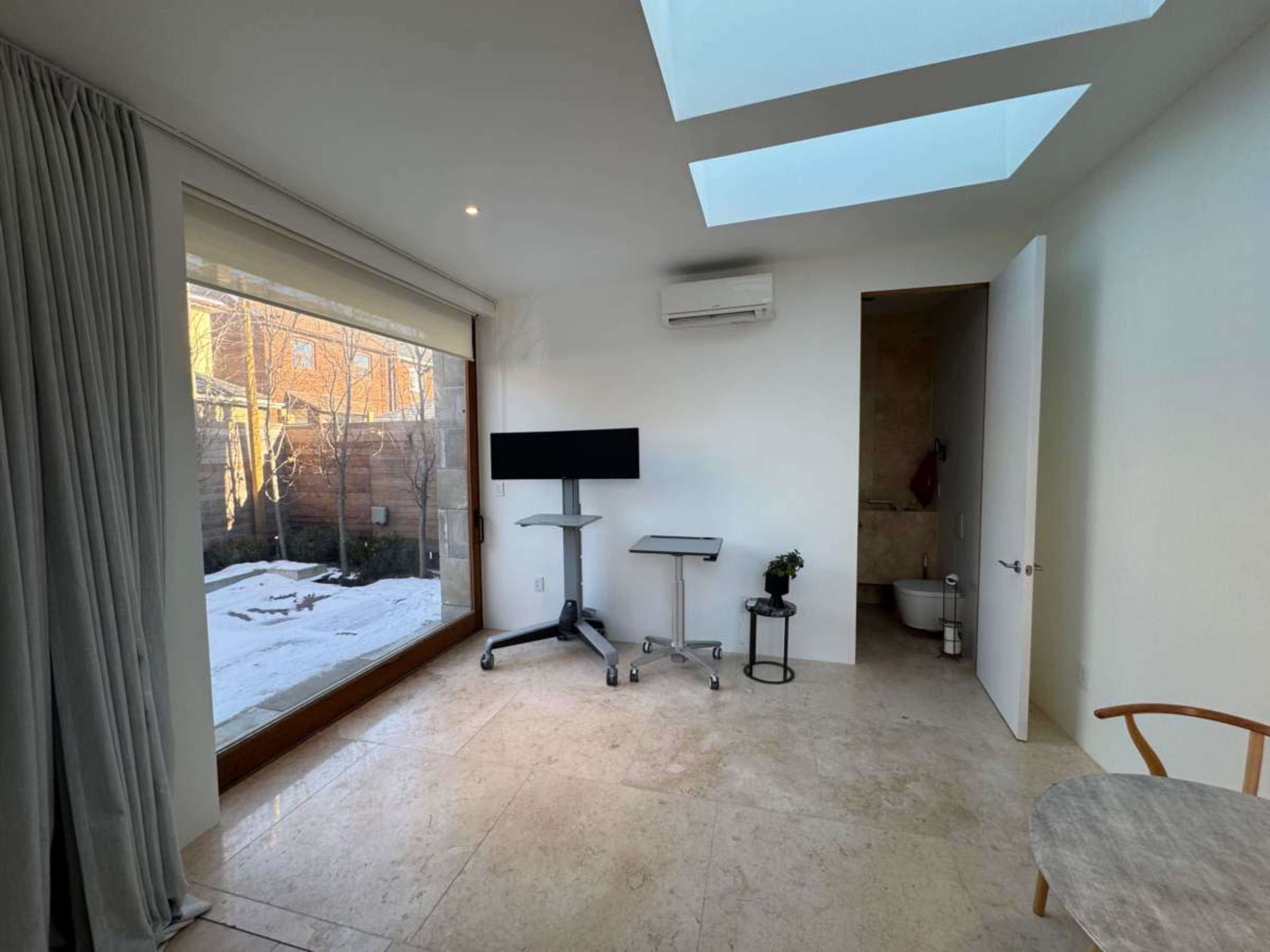 The image shows a minimalist room with a large window overlooking a snowy outdoor scene, featuring a small workspace with a monitor and a bathroom door in the corner.