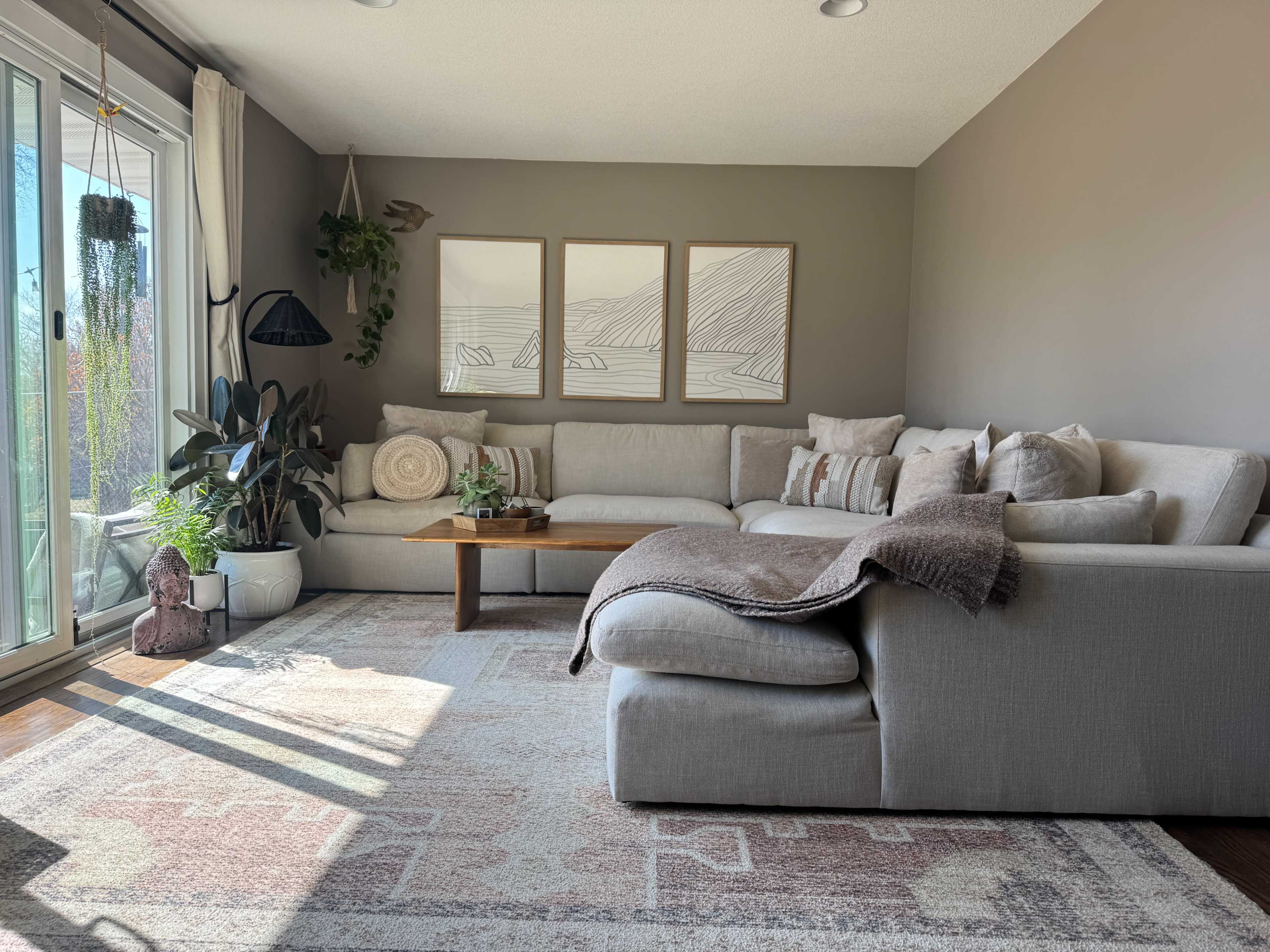 A modern living room features a large sectional sofa, a wooden coffee table, and framed artwork on the wall, with plants and natural light streaming in from a nearby window.