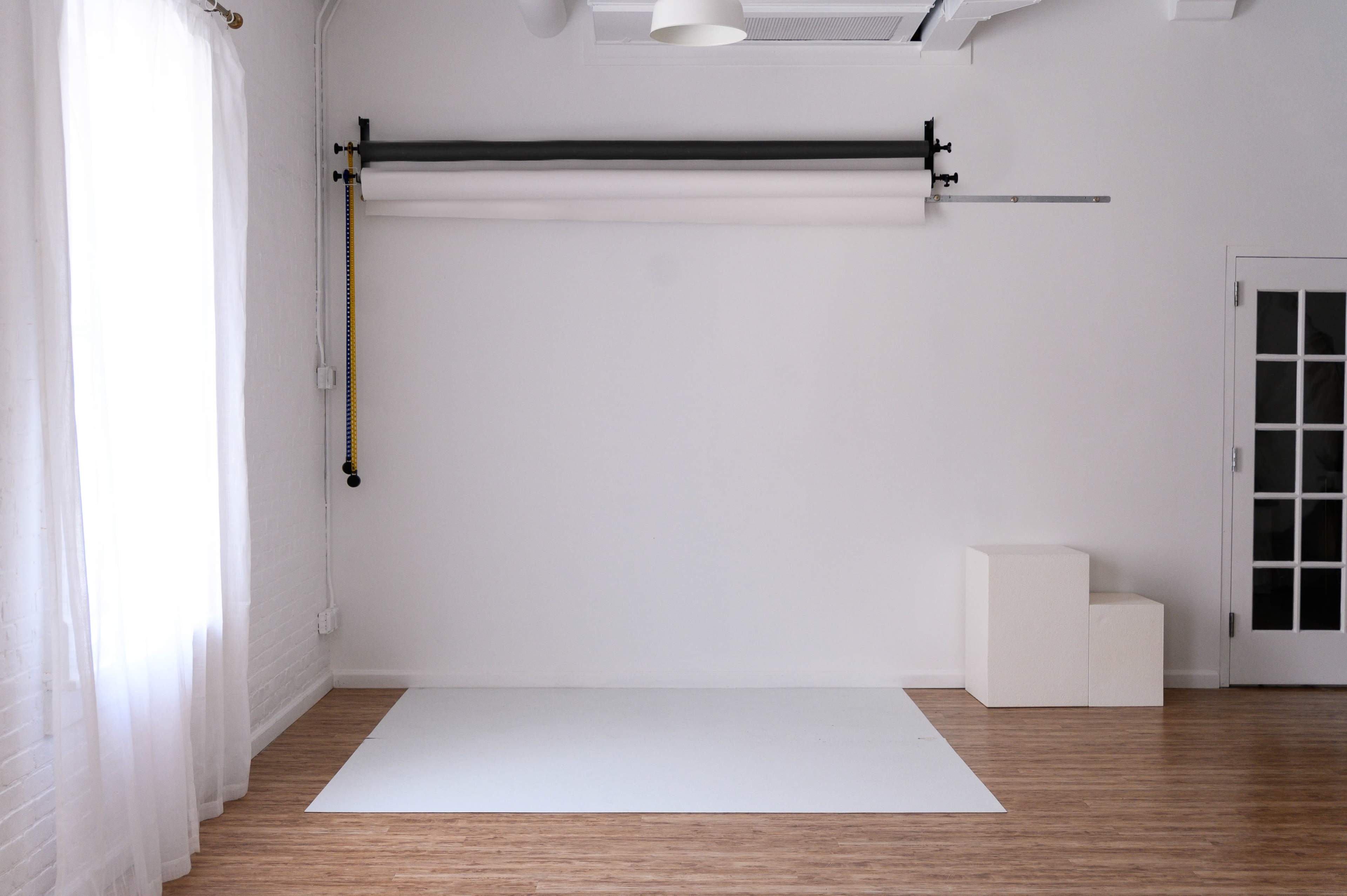 A bright, minimalist studio space with a white wall, wooden flooring, and a backdrop setup along with a small set of white cubes.