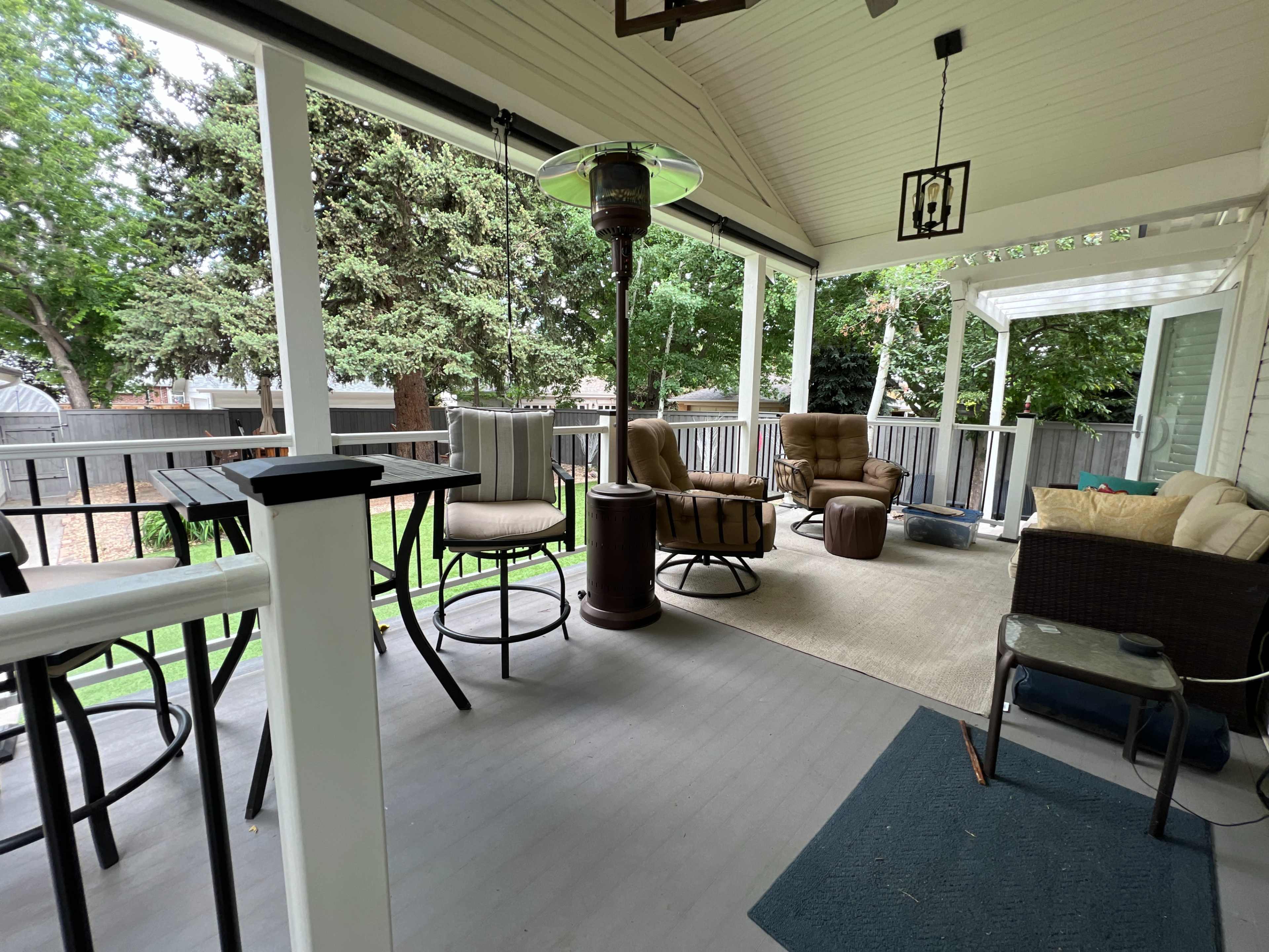 The image shows a covered patio featuring various seating options, including a pair of brown reclining chairs, a table with high stools, and a beige rug, surrounded by greenery.