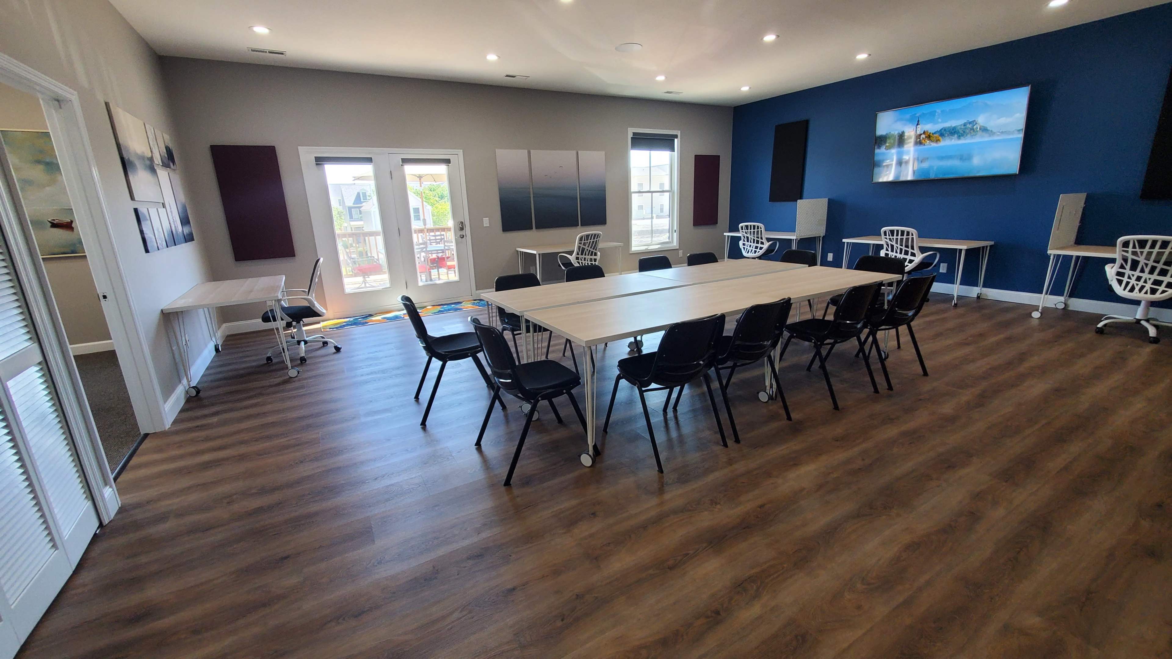 The image shows a modern conference room with a large table surrounded by black chairs, two screens mounted on the walls, and large glass doors opening to a view outside.