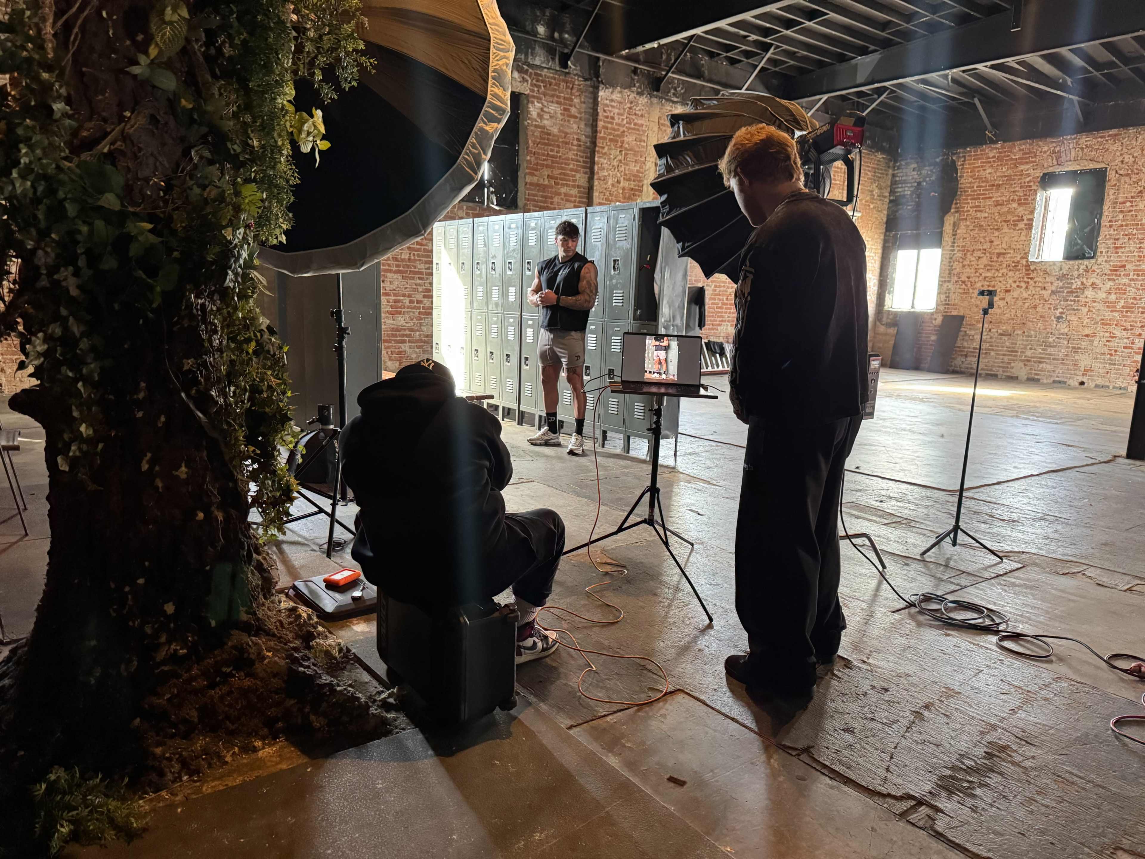 A photographer is setting up a shoot with a model in front of lockers, while another person sits on a case beside a tree inside a spacious industrial room.