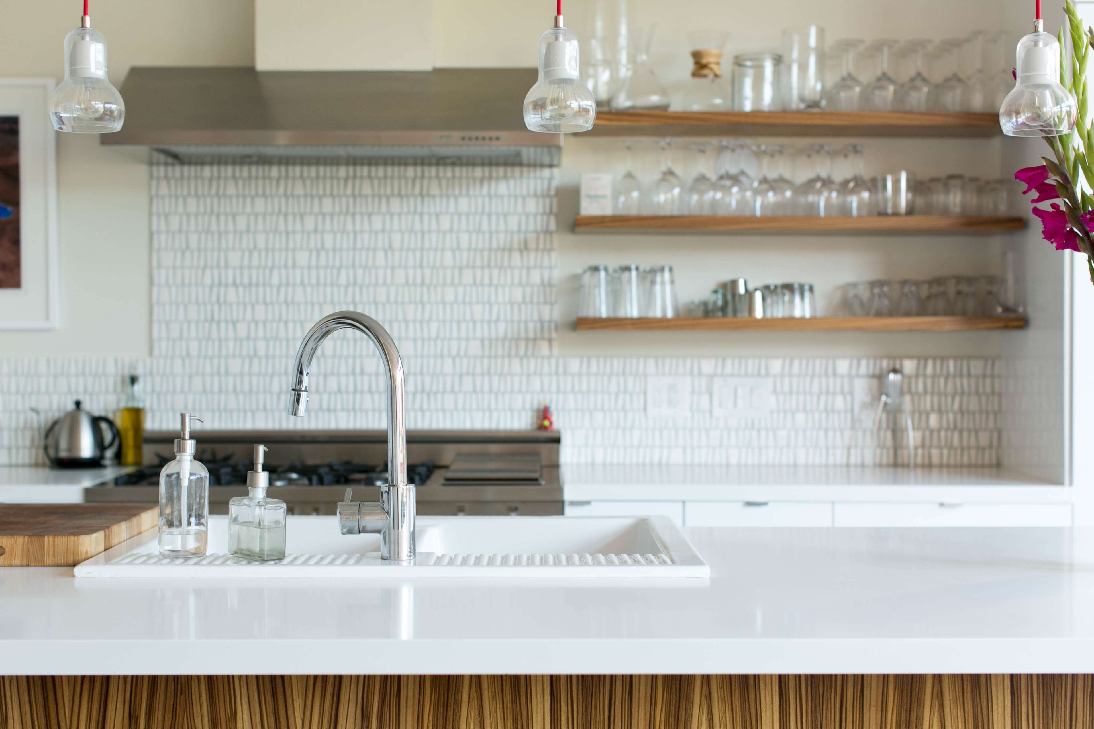 A modern kitchen features a white countertop with a sink, glass storage shelves, and a stainless steel stove in the background.