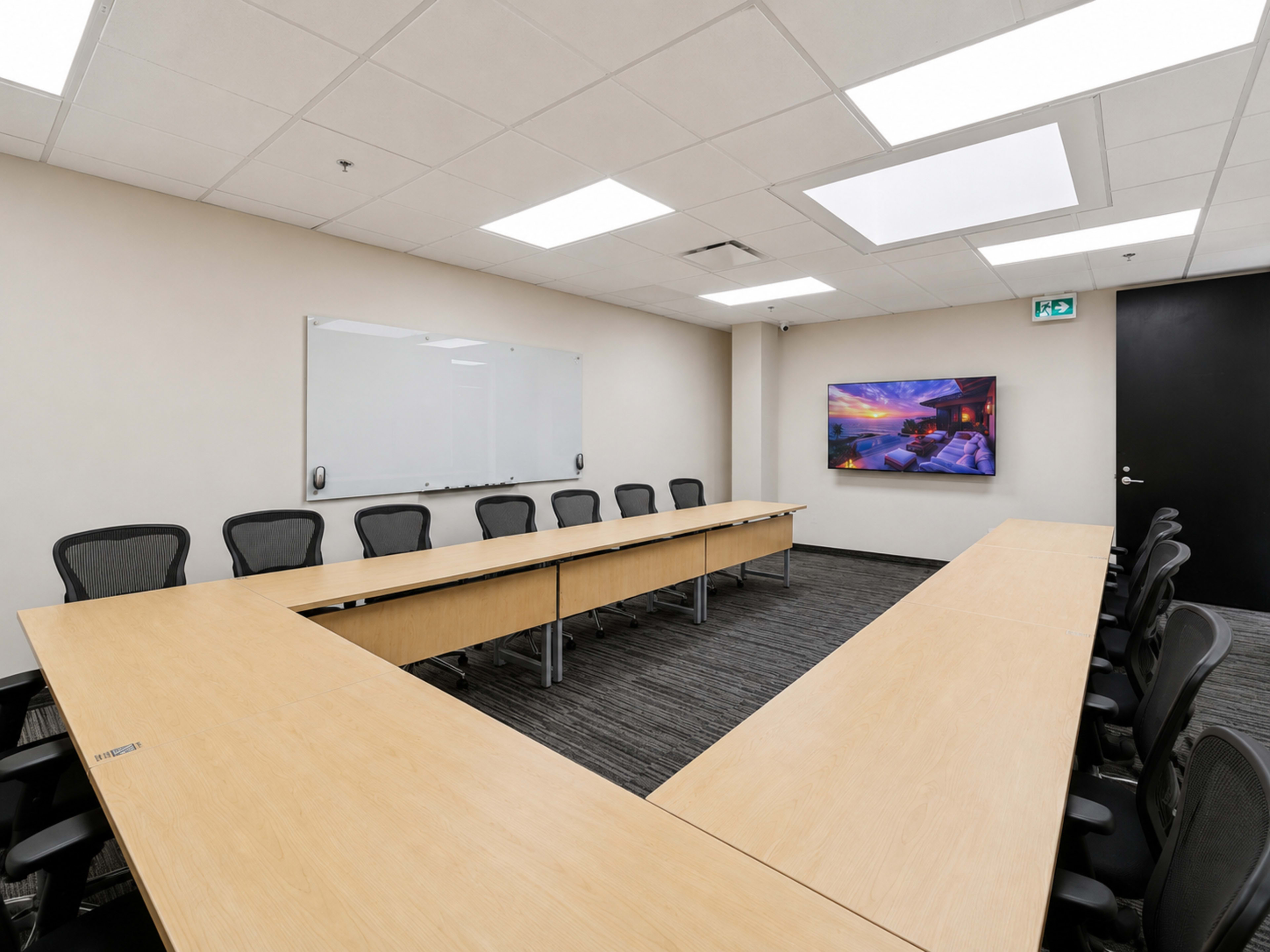 A large conference room features a long wooden table arranged in a U-shape, surrounded by black office chairs, with a whiteboard and a television screen on the walls.