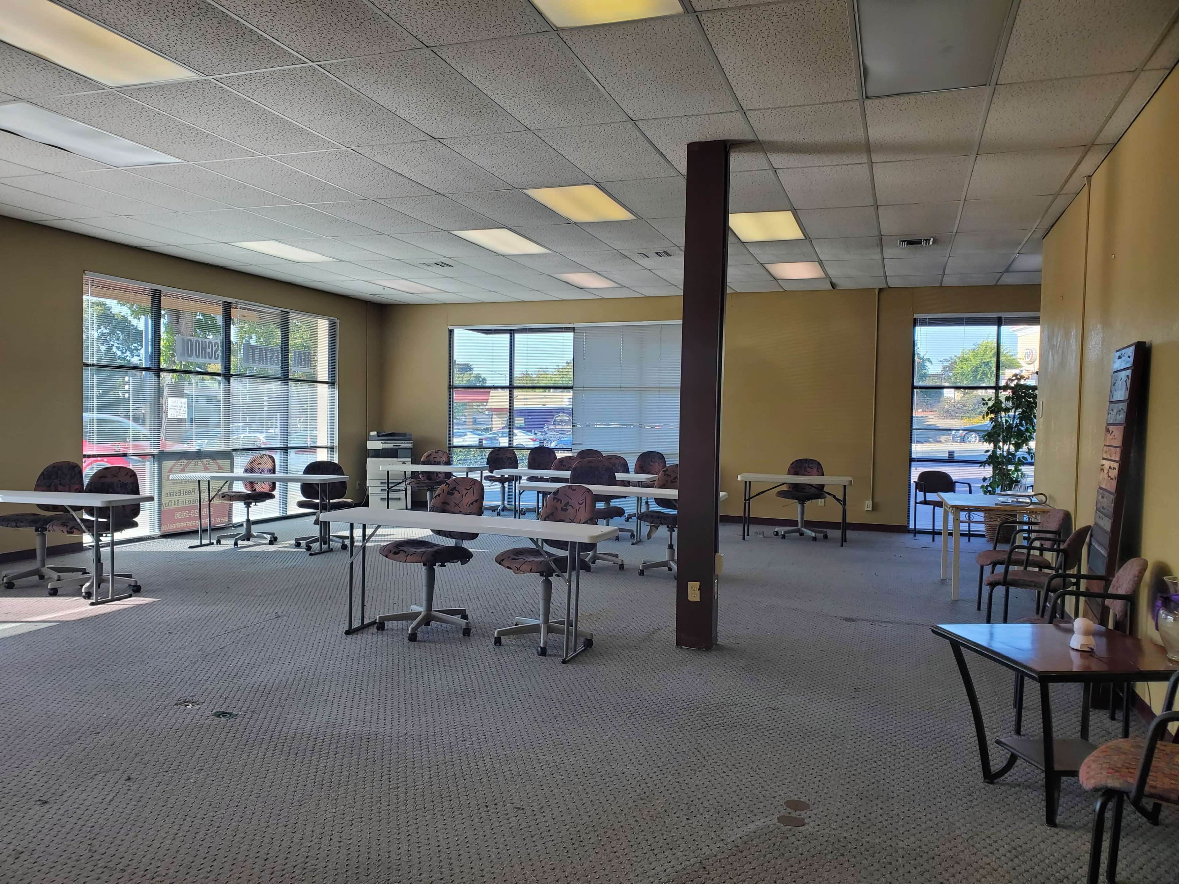 An empty room with several tables and chairs arranged throughout, large windows on one side letting in natural light, and a carpeted floor.