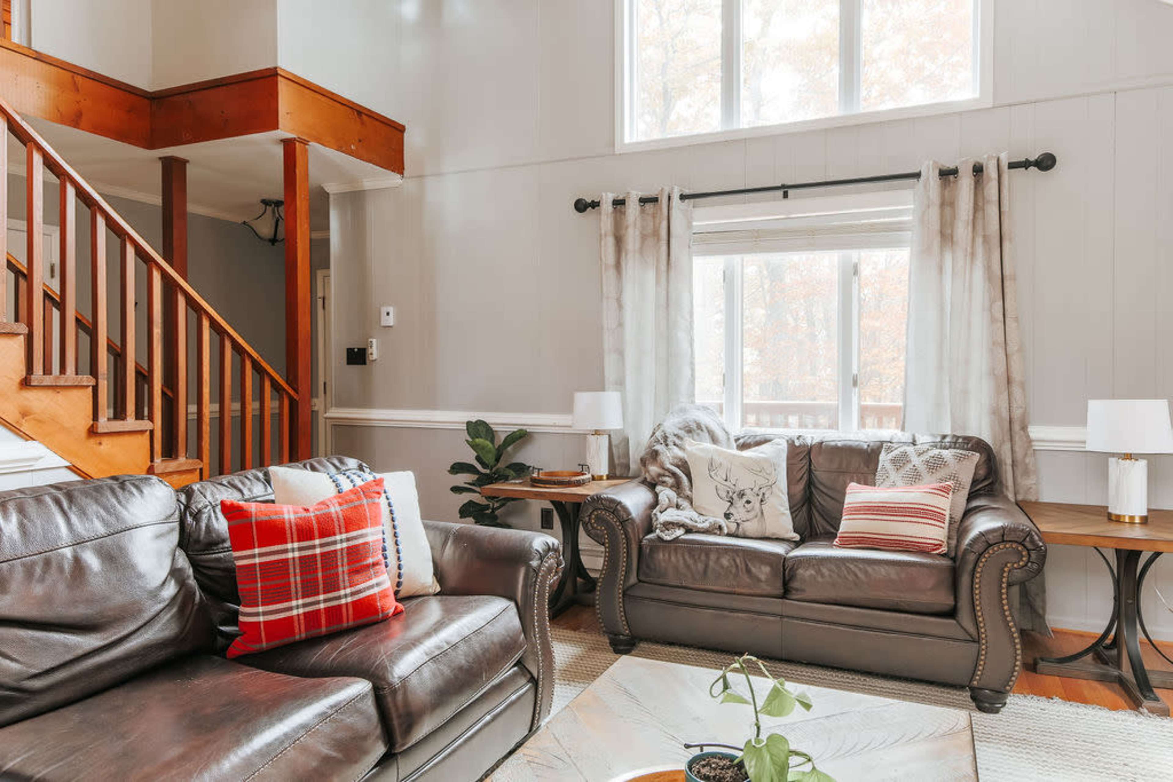 The living room features two leather sofas, a wooden staircase, and large windows with sheer curtains allowing natural light.