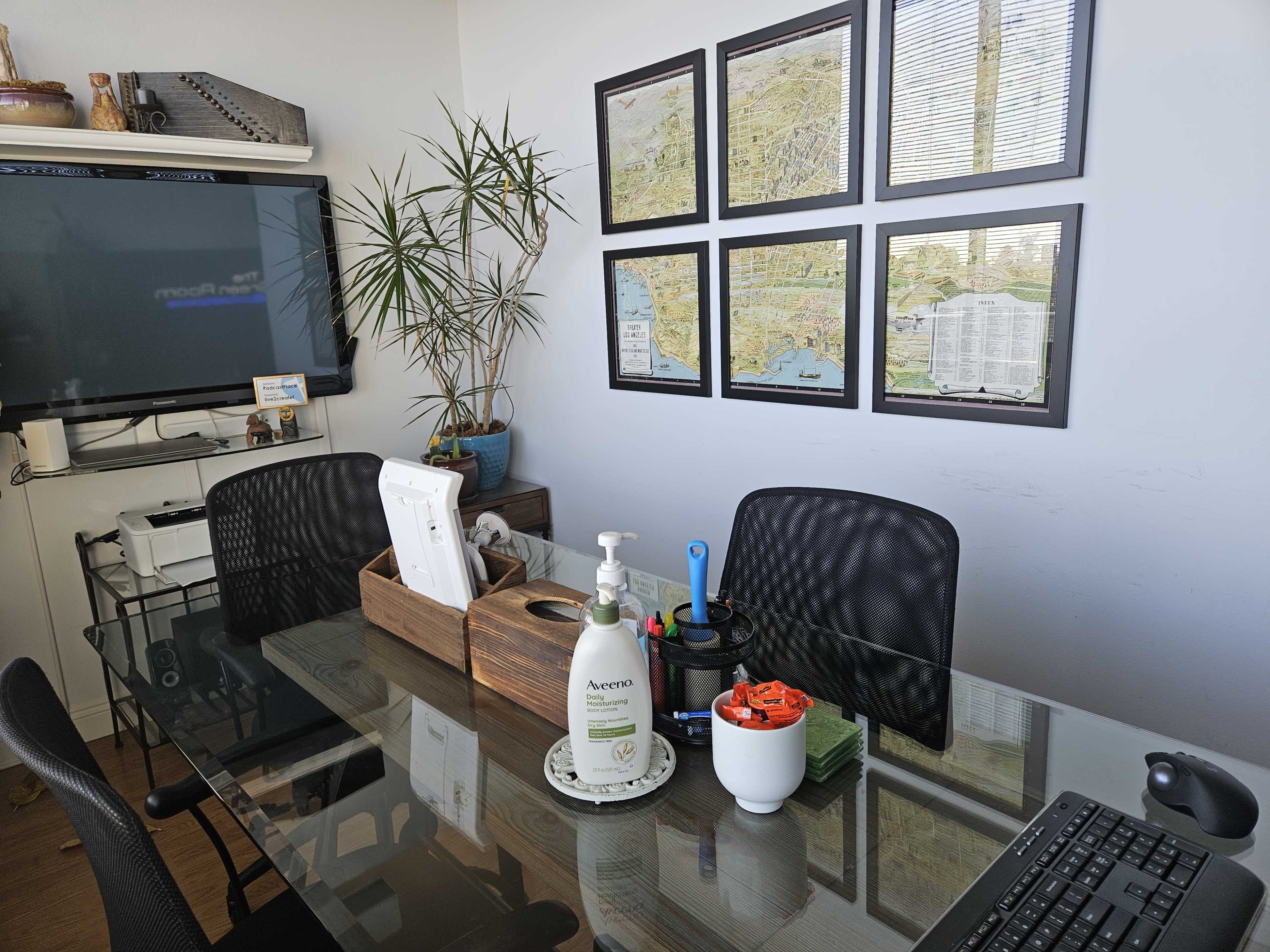 A modern office space featuring a glass desk, a TV on the wall, potted plants, and framed maps.