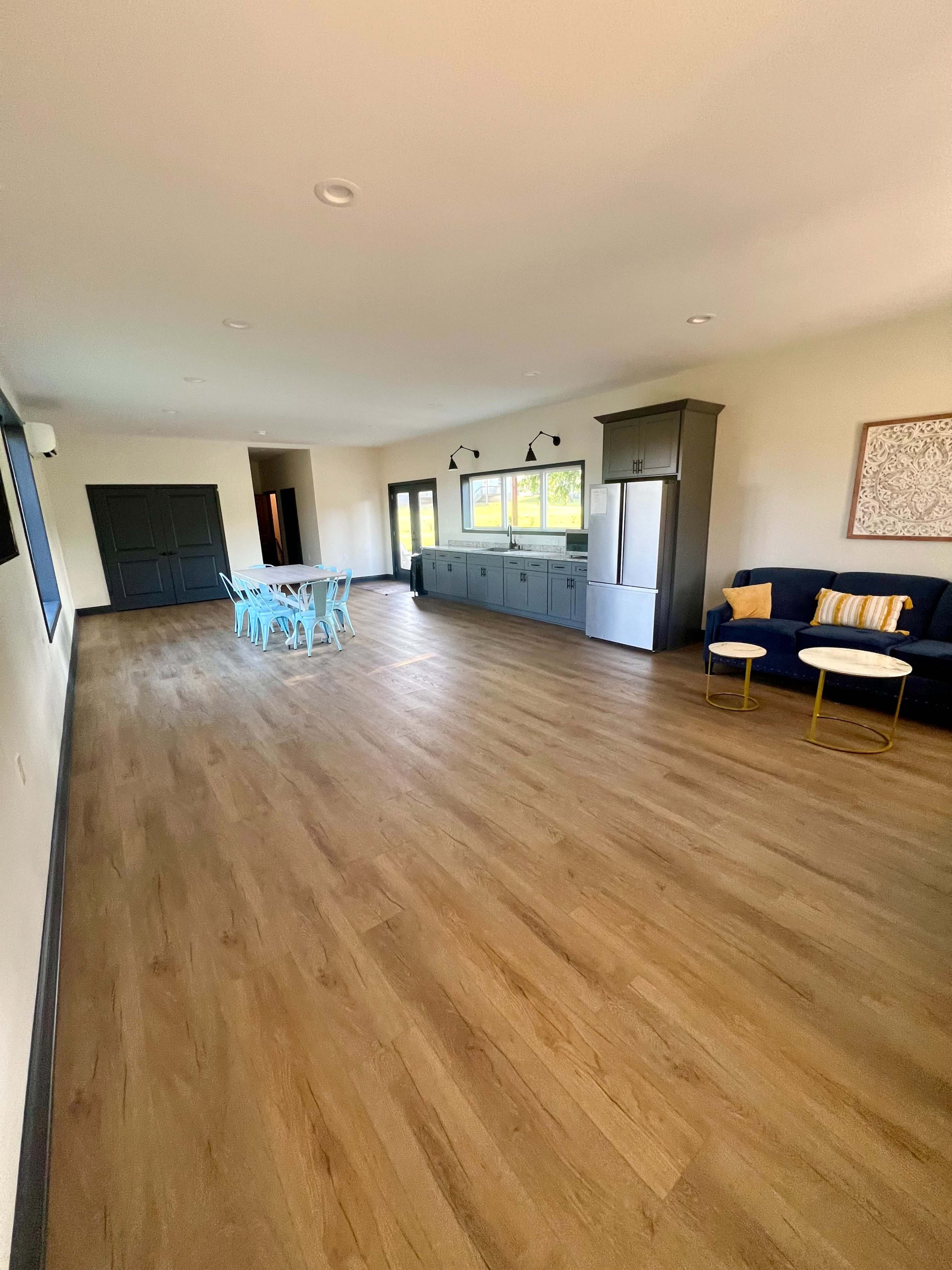 The image shows a spacious room with light wooden flooring, a kitchen area with gray cabinets, a refrigerator, a dining table with blue chairs, and a small seating area with a dark sofa and side tables.