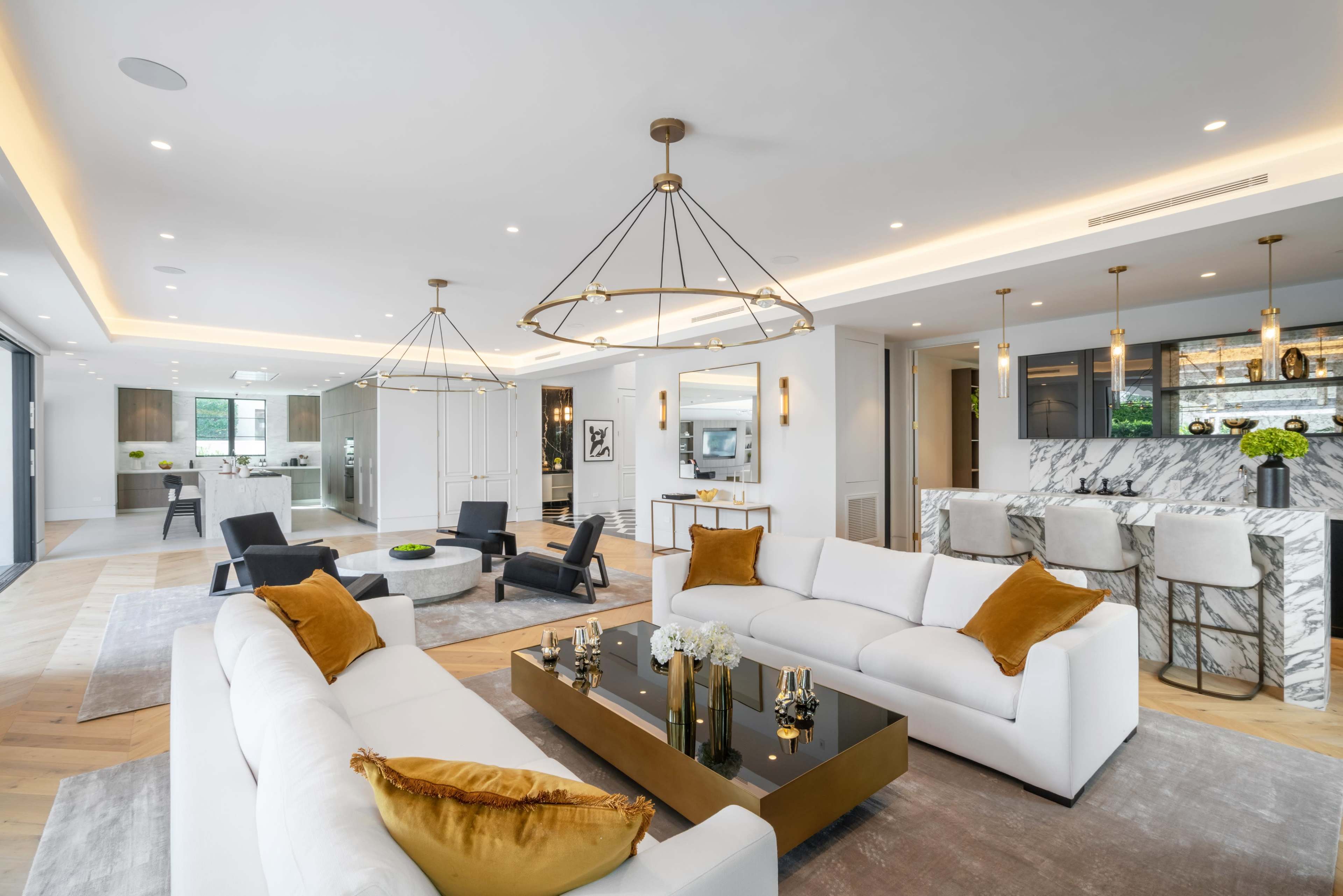 A modern living room features two white sofas, a black coffee table, and a kitchen area in the background with marble countertops and pendant lighting.