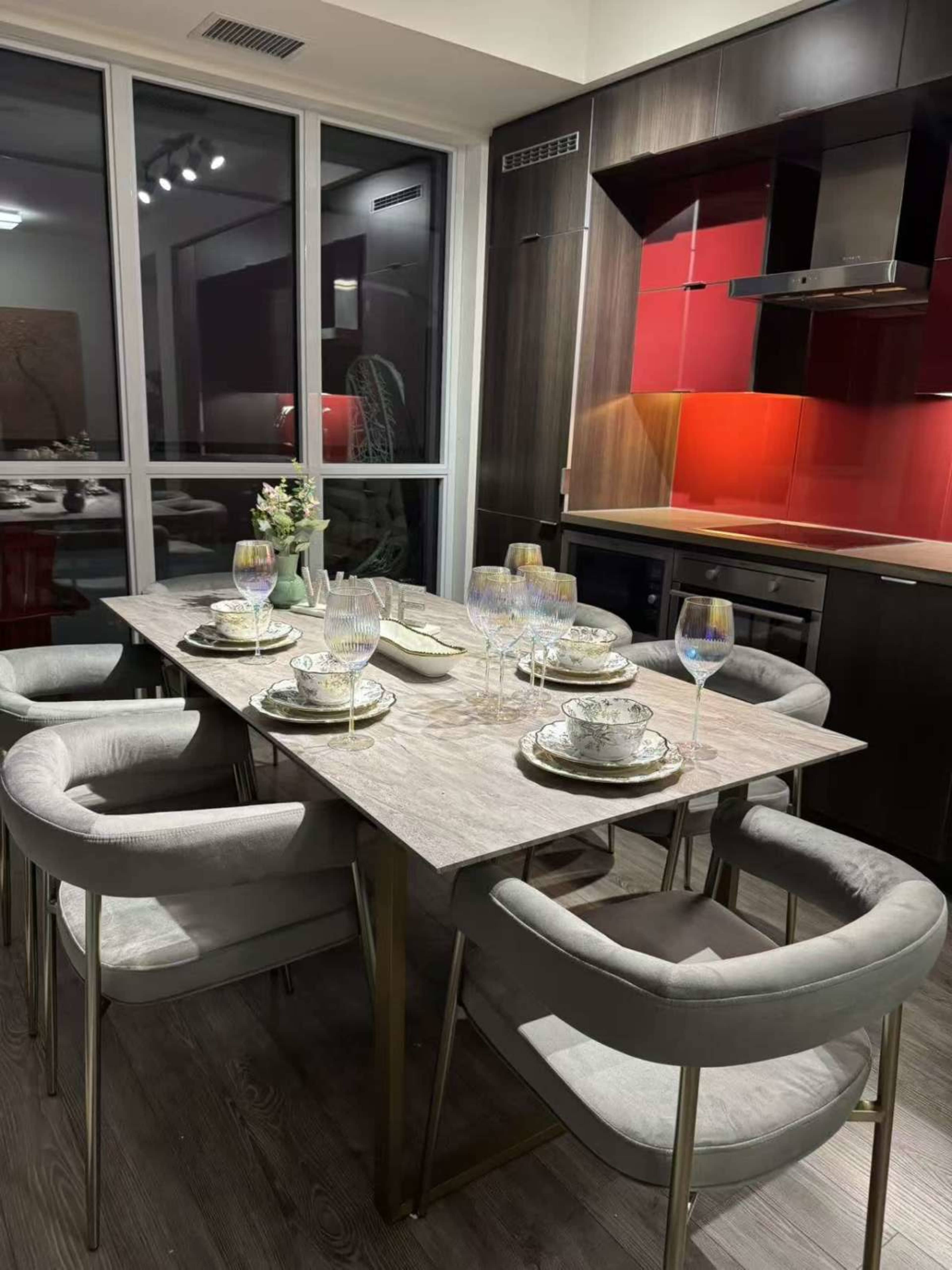 A dining table is set with elegant dishware and wine glasses in a modern kitchen with red and dark wood cabinetry.