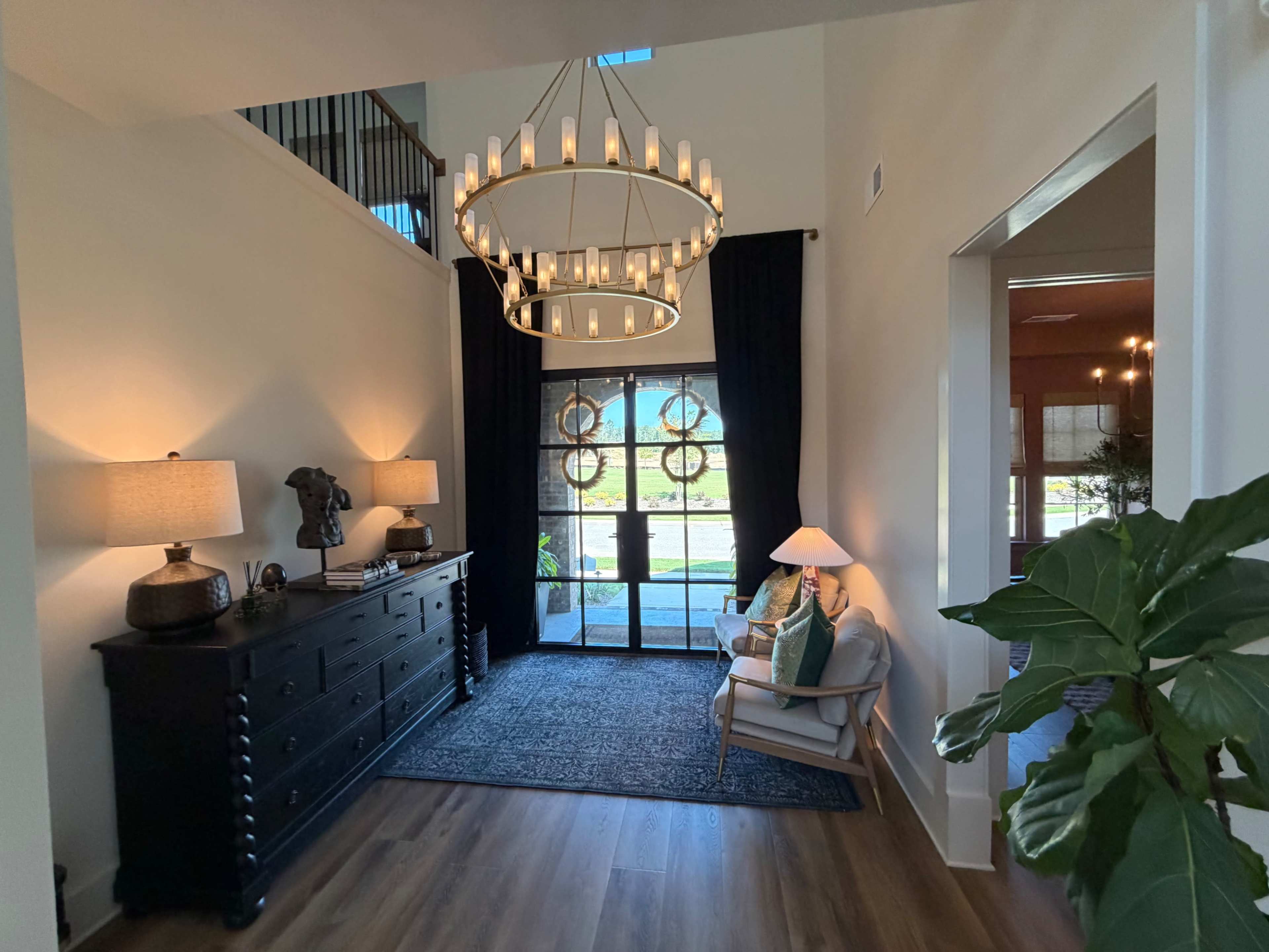 A spacious entryway features a large chandelier, two lamps on a dark dresser, and a seating area beside a large window with decorative circular mirrors.