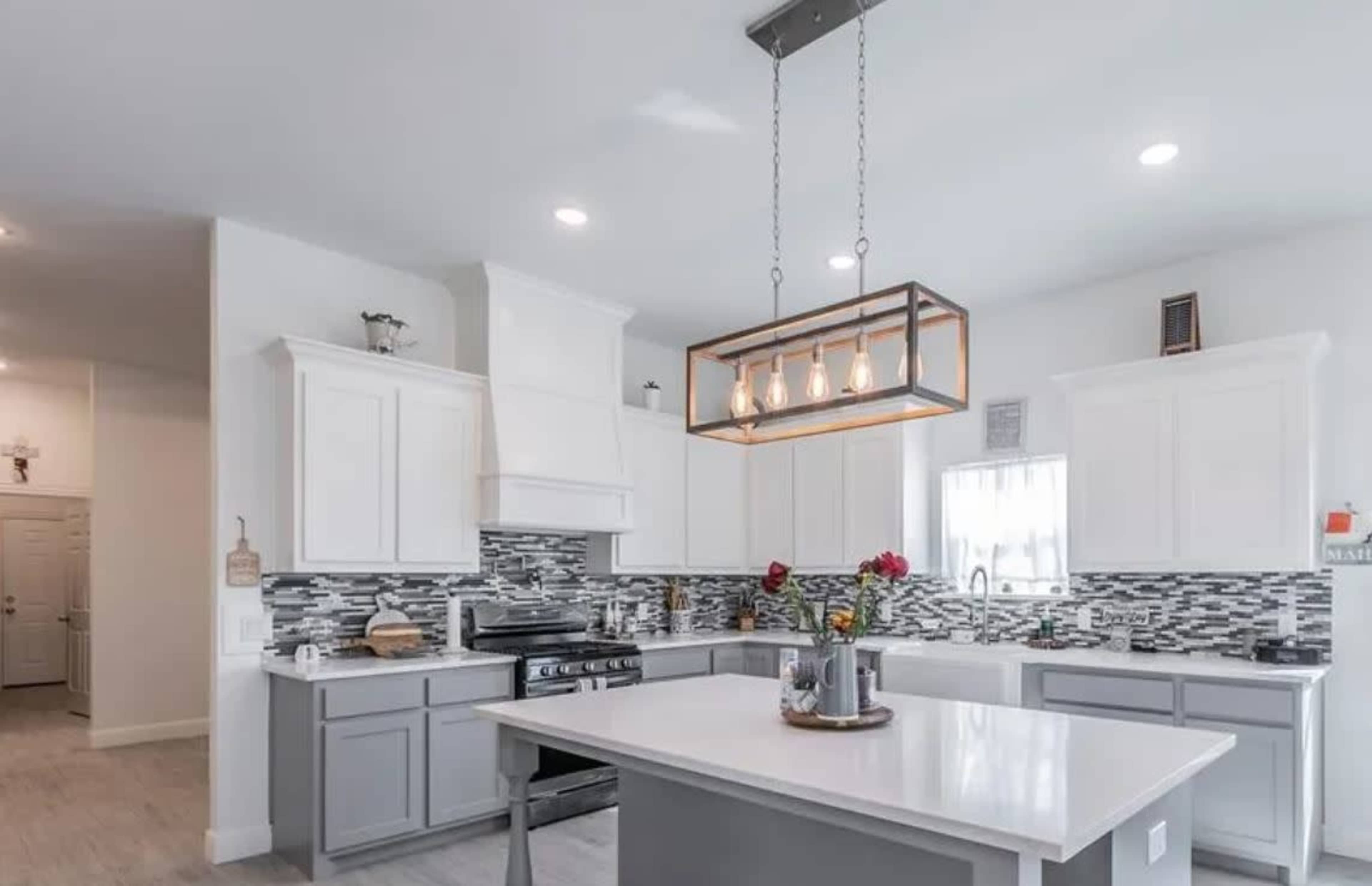 A modern kitchen features white cabinetry, gray lower cabinets, a central island with a stovetop, and a decorative light fixture above.