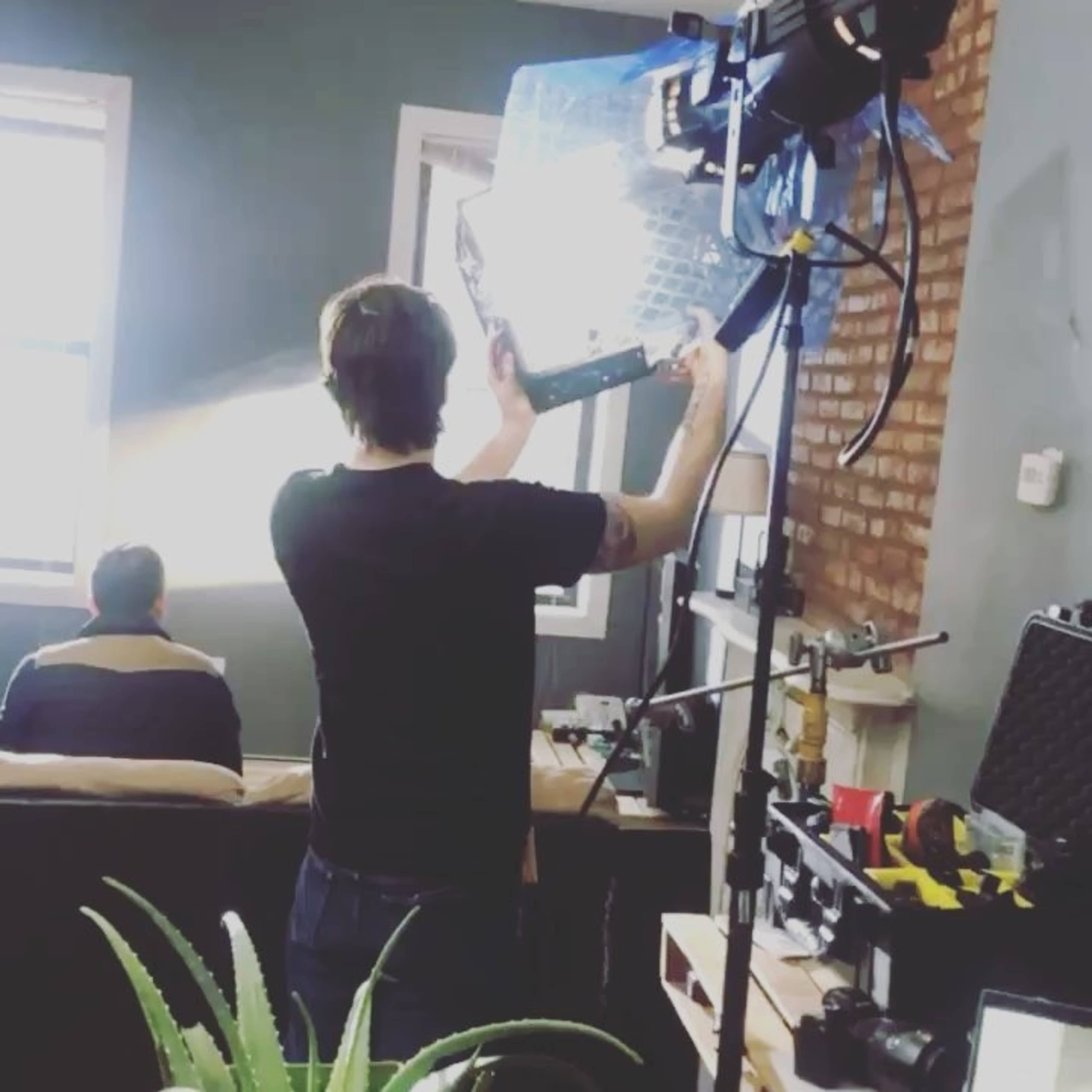 A person is adjusting a light reflector while another individual sits on a couch in a well-lit room.