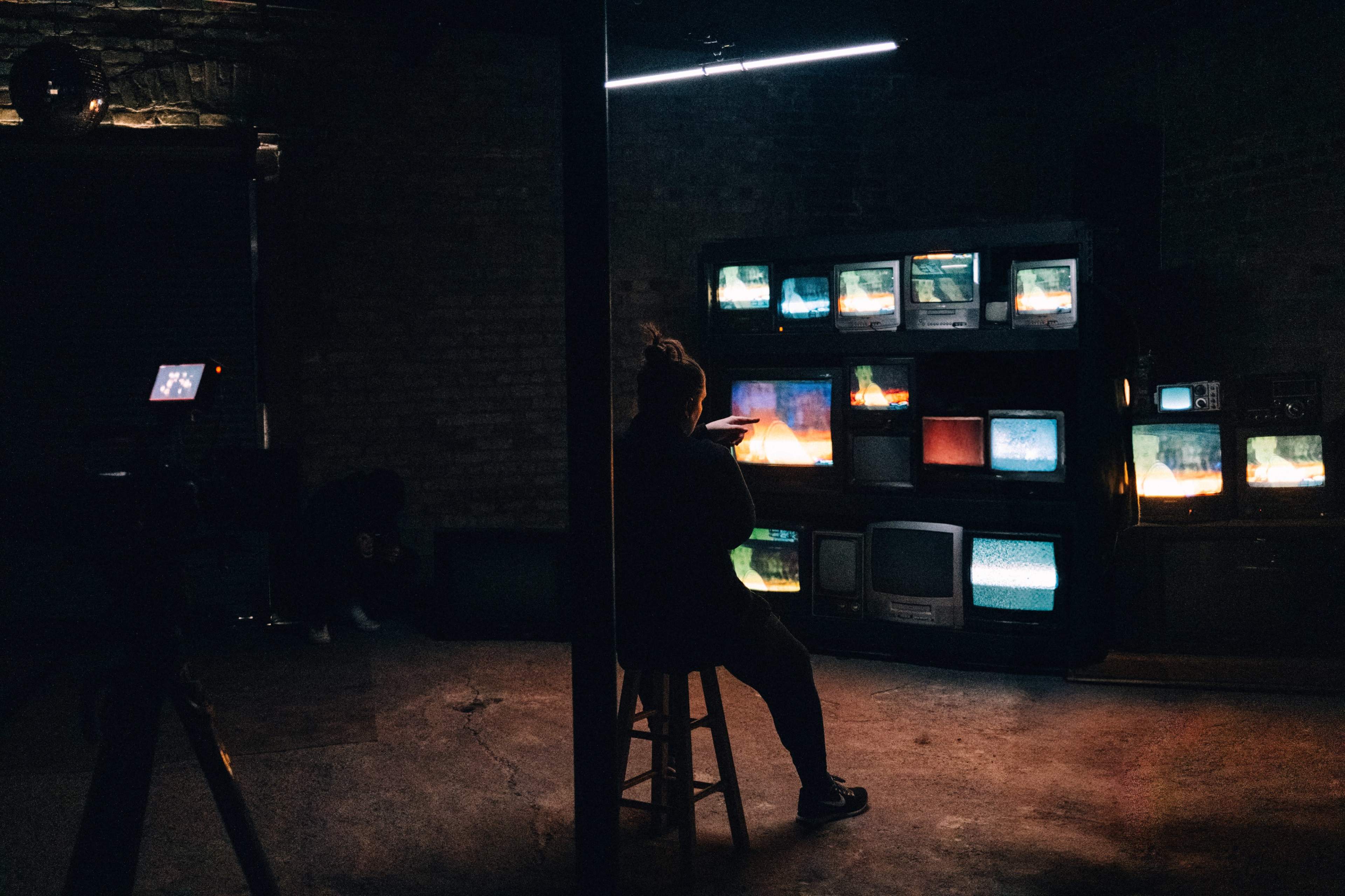 A person sits on a stool in a dimly lit room surrounded by multiple vintage televisions that display various static images.