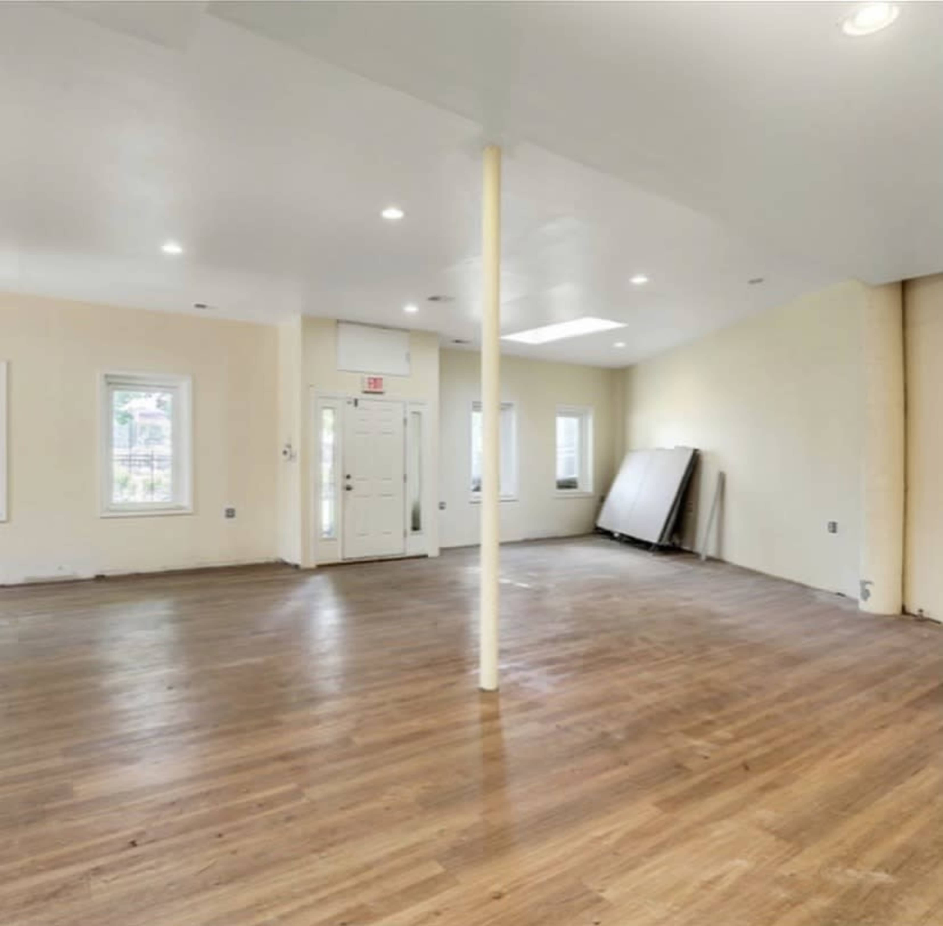 The image shows a spacious, empty room with wooden flooring, a central support column, and windows providing natural light.