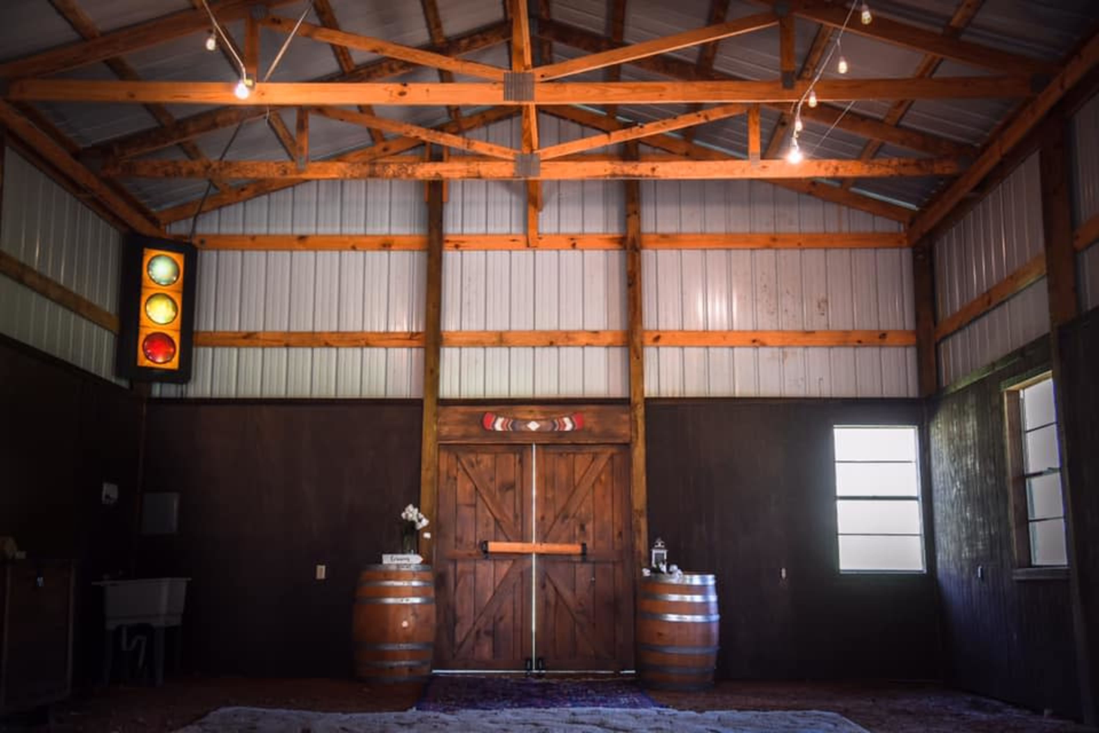 The interior of a barn features wooden beams, two large barrels, a traffic light, and windows on either side of a central wooden door.