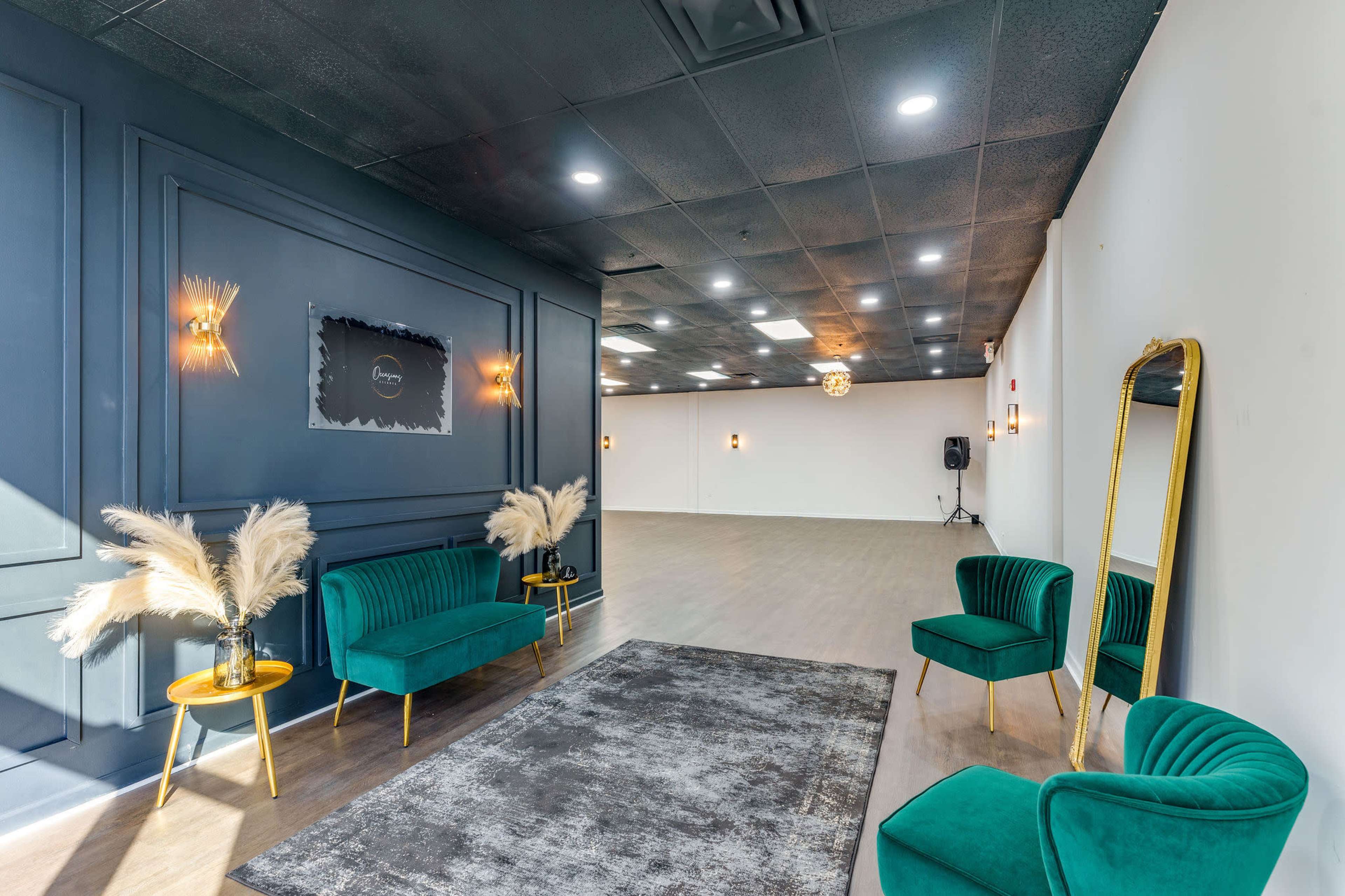 A spacious interior with dark walls, two green velvet chairs, a large mirror, and decorative lighting fixtures, creating an inviting atmosphere.