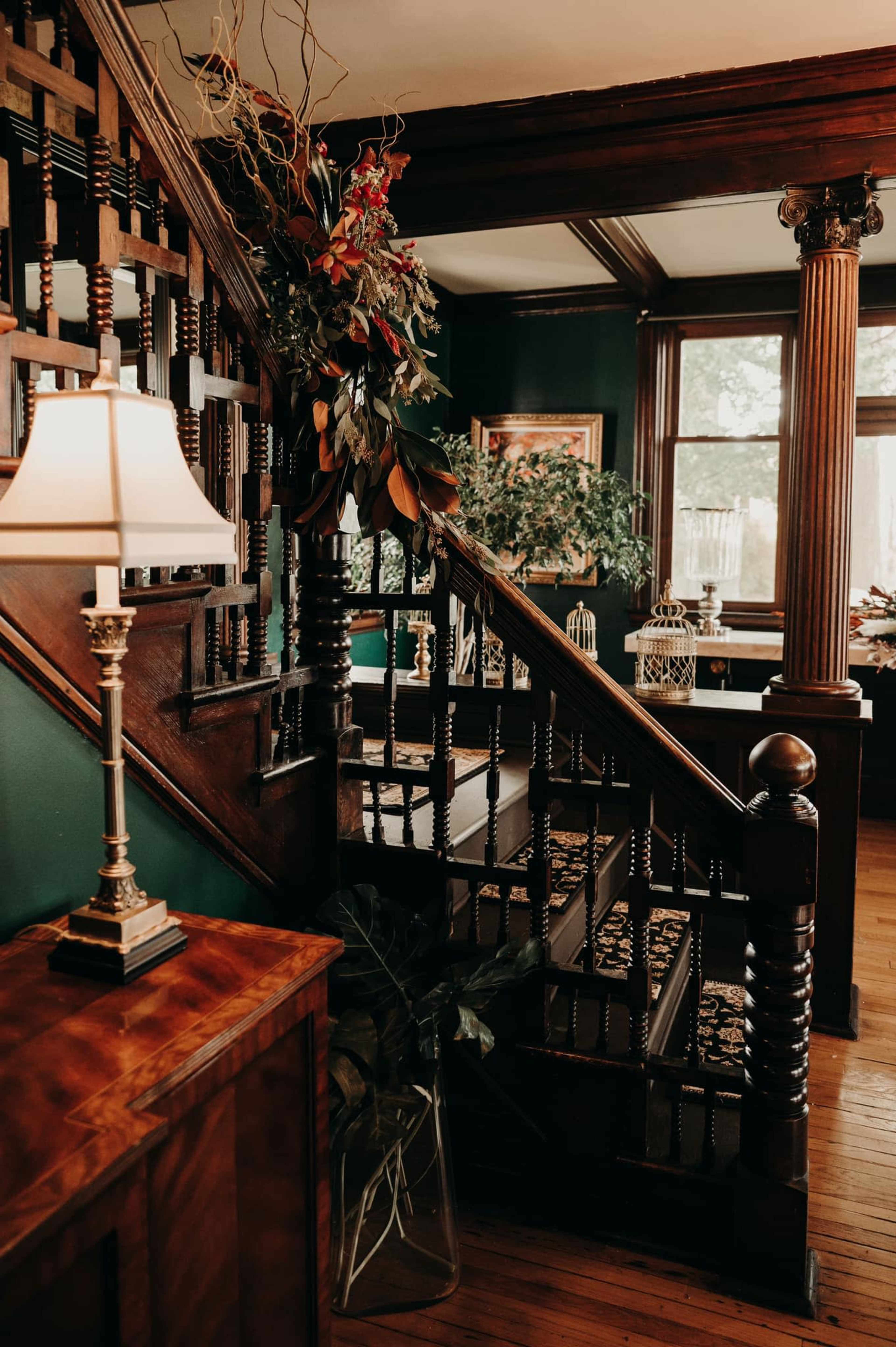 A staircase with ornate wooden railings, surrounded by potted plants and a lamp, set against a dark green wall in a well-lit room.