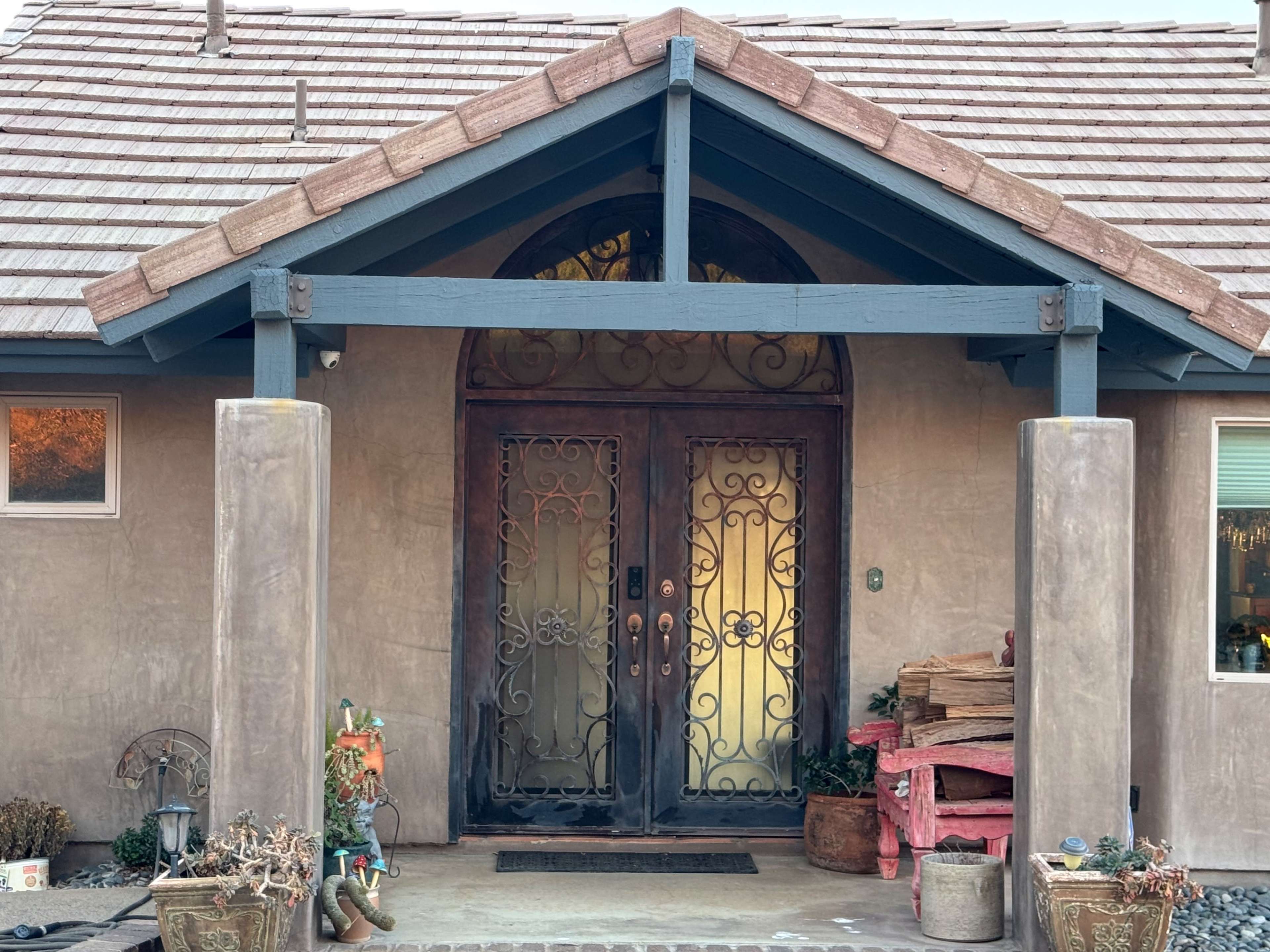 The image shows the entrance of a home, featuring a double door with decorative wrought iron designs, flanked by two columns under a pitched roof.