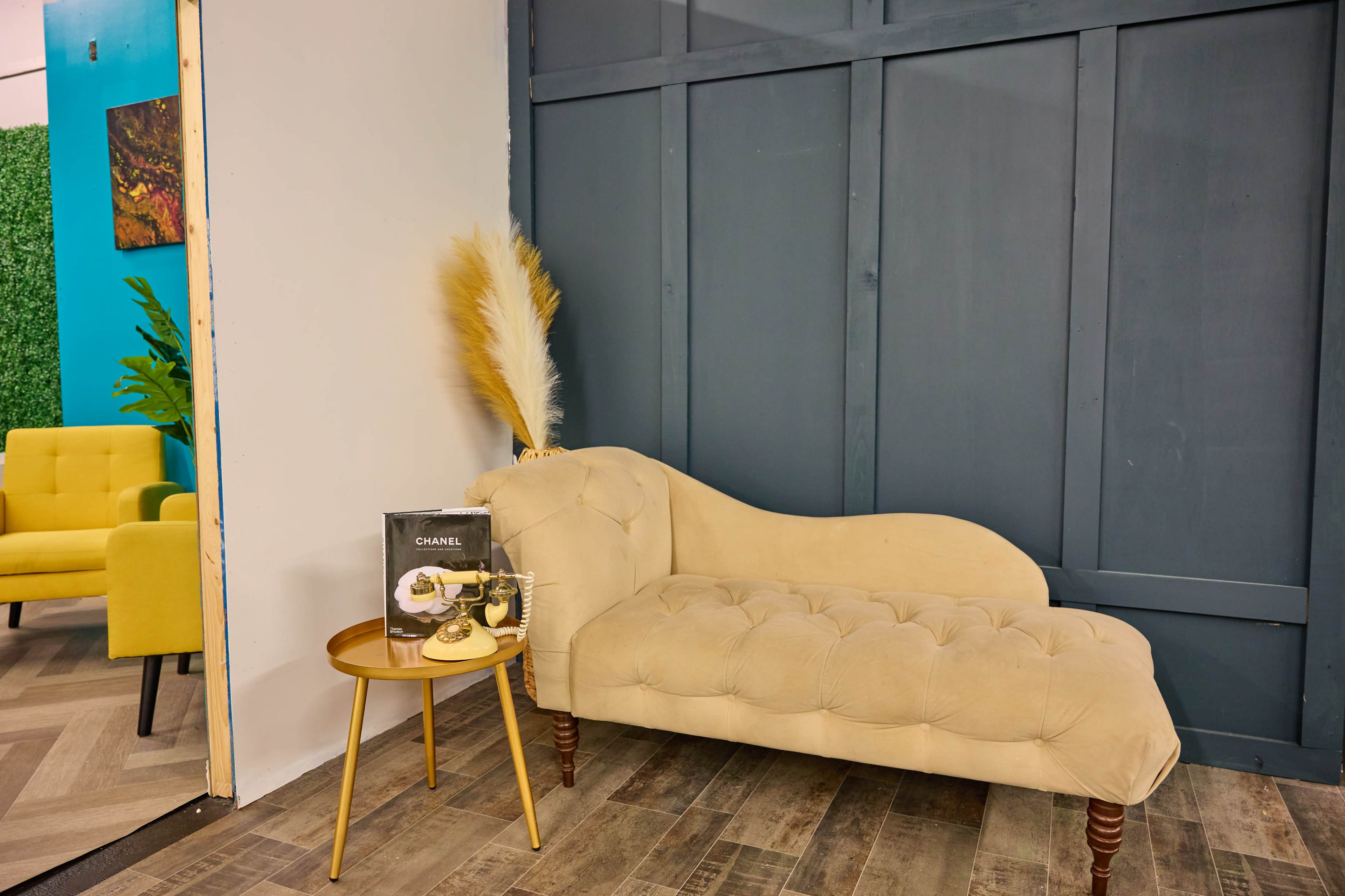 A tufted beige chaise lounge alongside a small gold side table with styled magazines and décor, set against a gray wall.