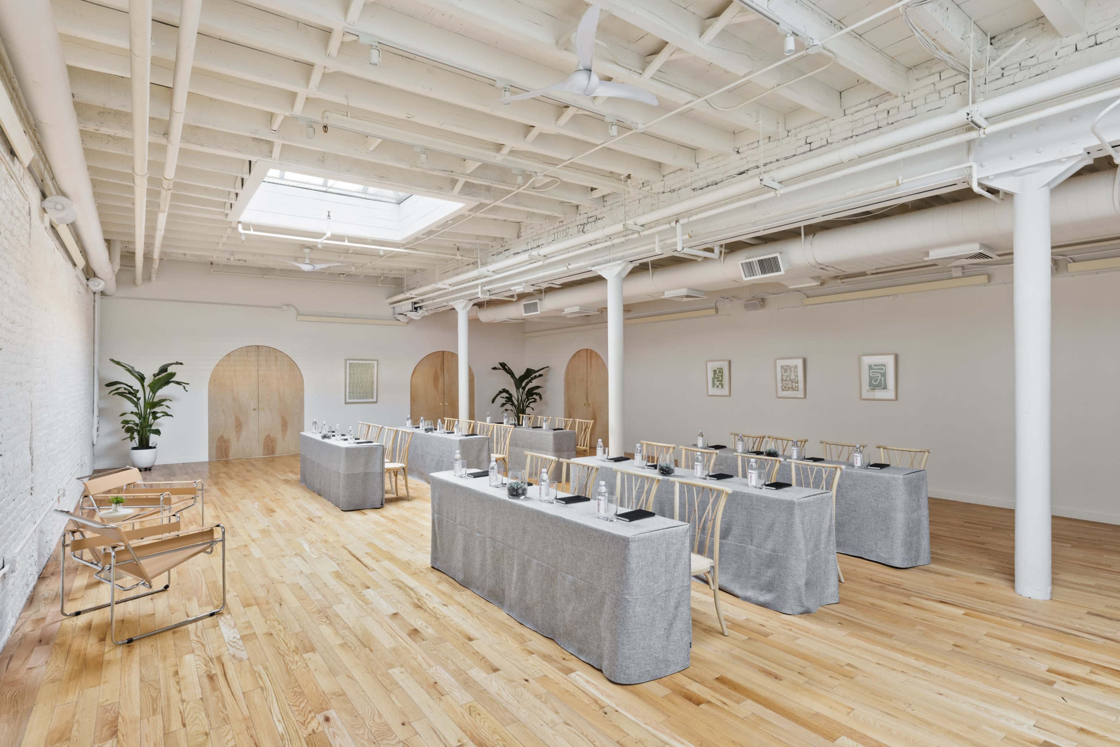 The image shows a modern meeting space with long tables arranged in a U-shape, surrounded by light wooden flooring and white walls, featuring plants and natural light from a skylight.