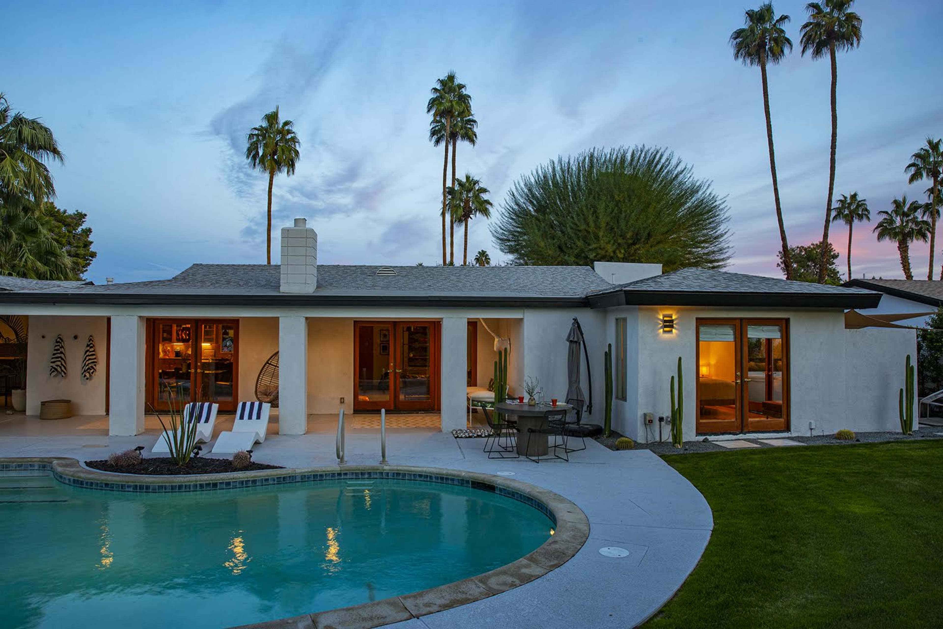 A modern single-story home with a pool and palm trees is set against a twilight sky.