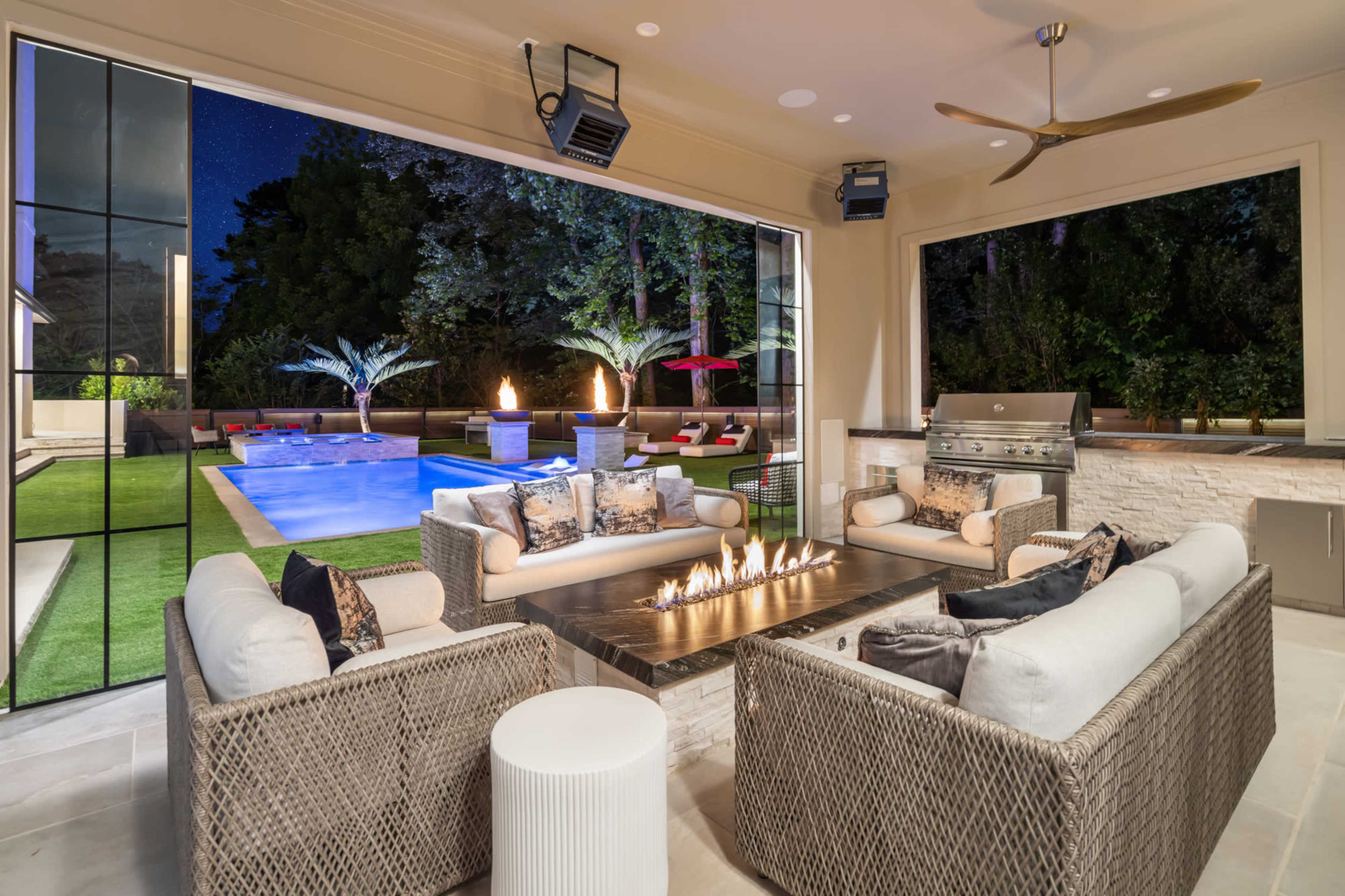The image shows a modern outdoor living space with a fire pit, comfortable seating, and a view of a pool surrounded by greenery and lounge chairs.