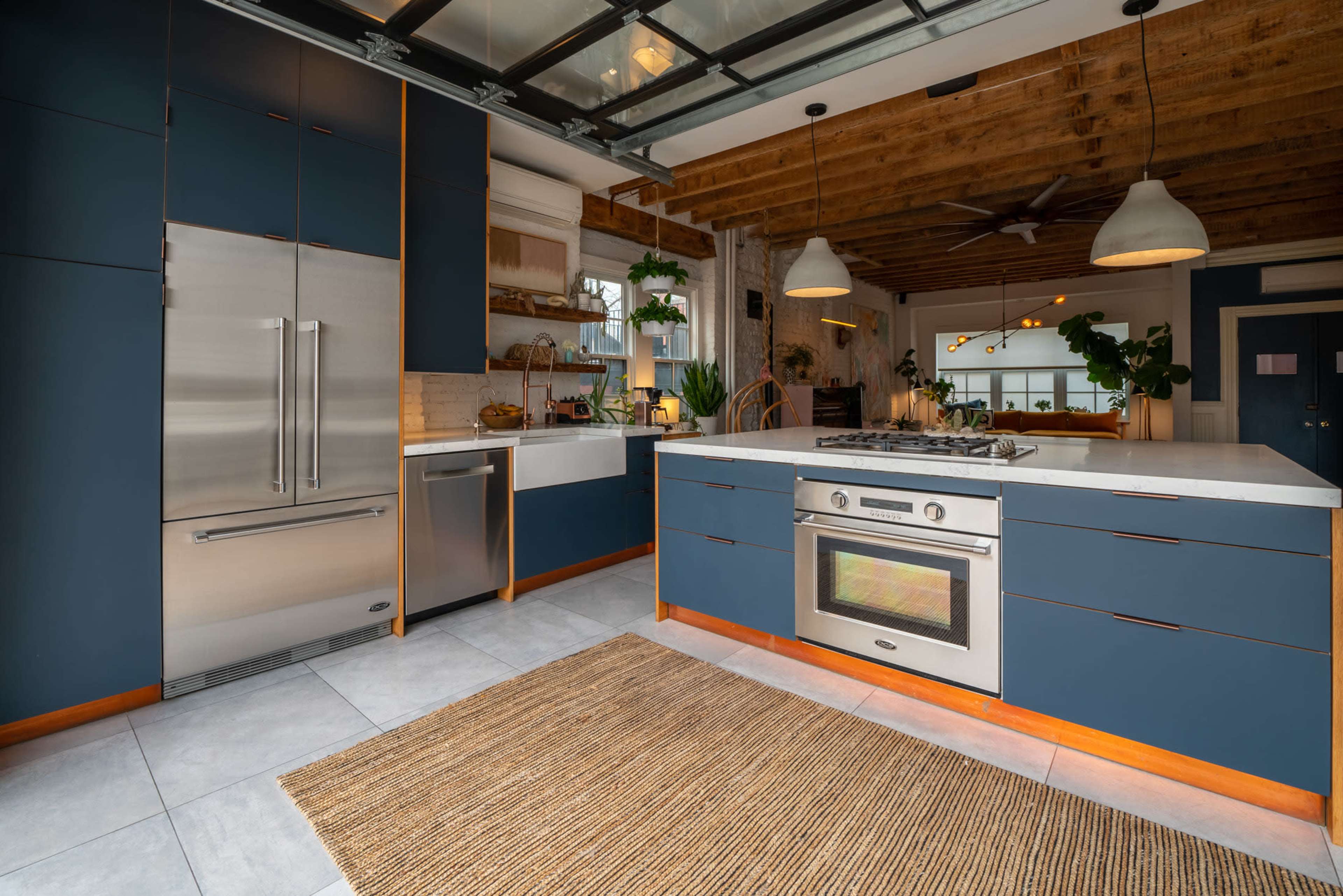 The image shows a modern kitchen with blue cabinets, a stainless steel refrigerator, a gas range in the center island, and wooden ceiling beams.