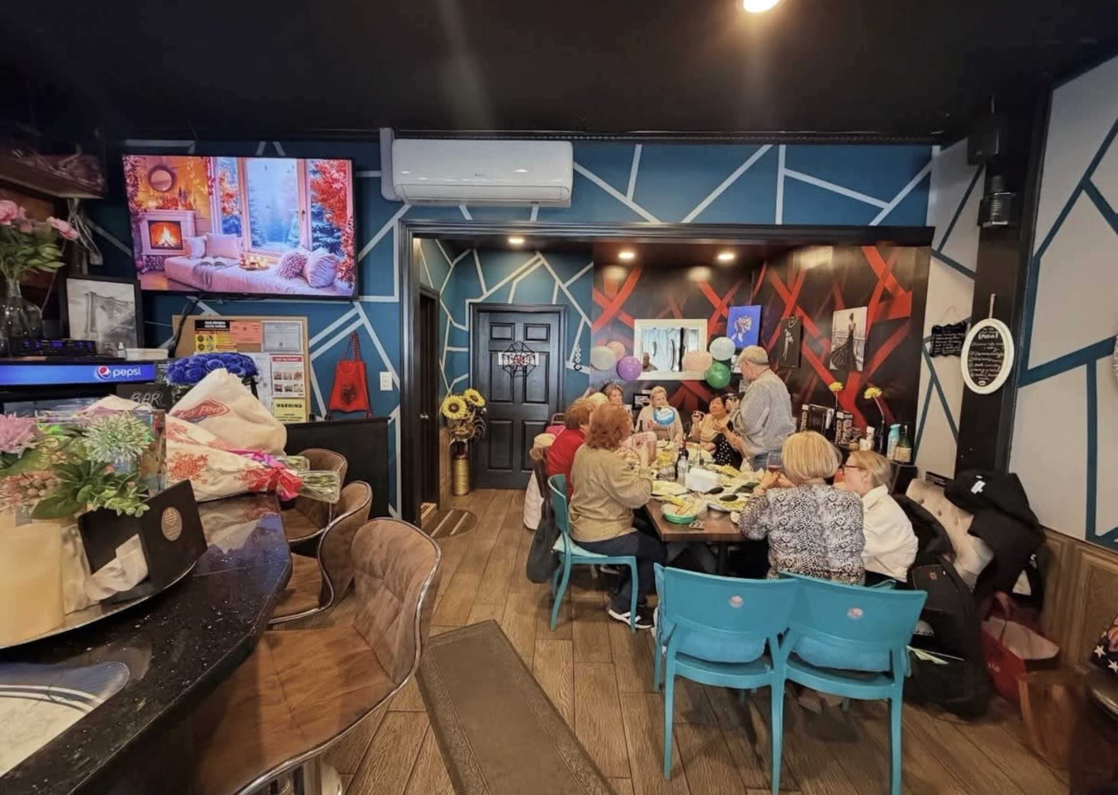 A group of people is gathered around a table in a vibrant café, while a television displays an image of a cozy scene on the wall.