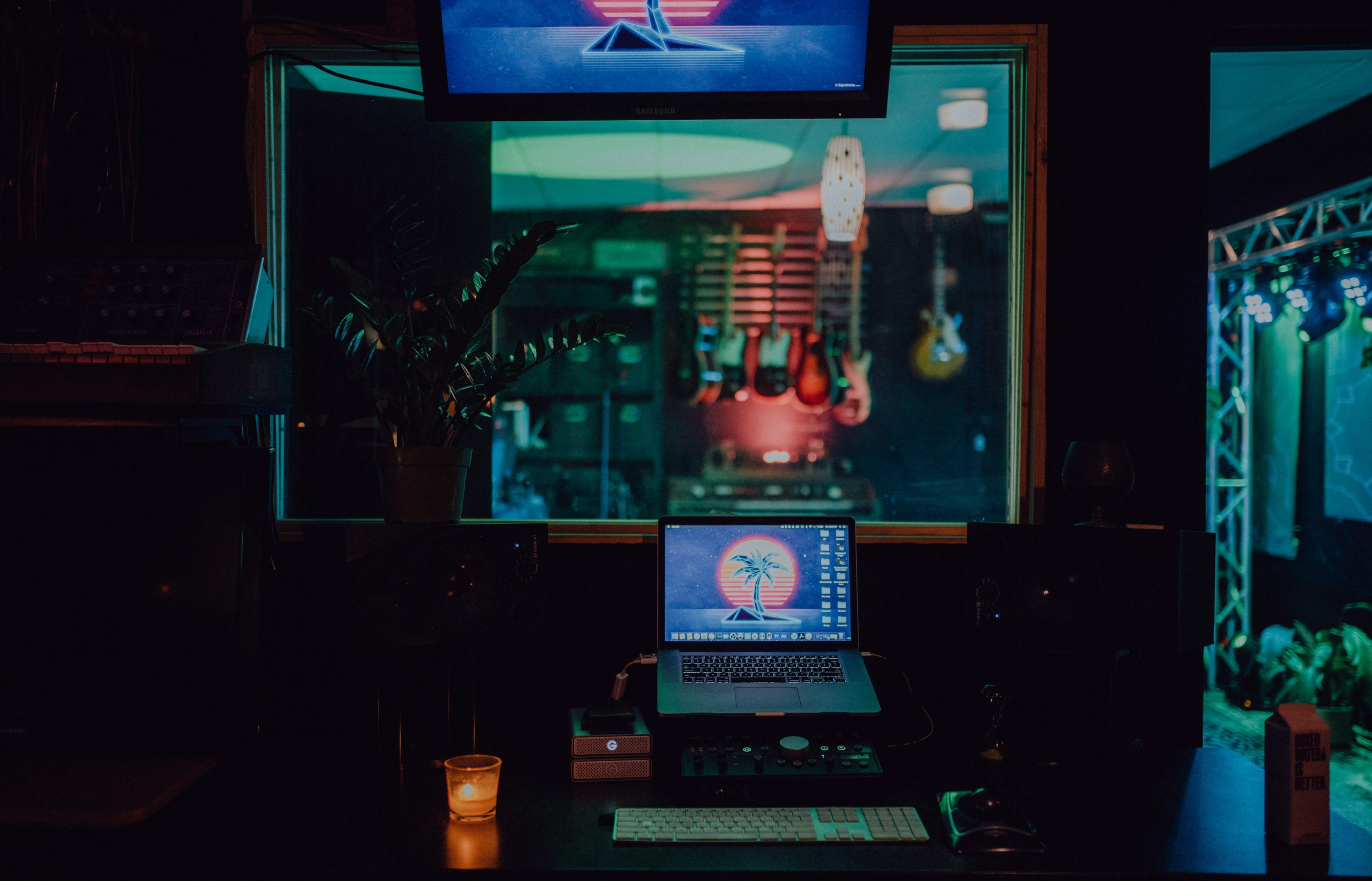 A dimly lit music studio features a laptop with a tropical wallpaper, surrounded by guitars and studio equipment, with colorful lighting in the background.