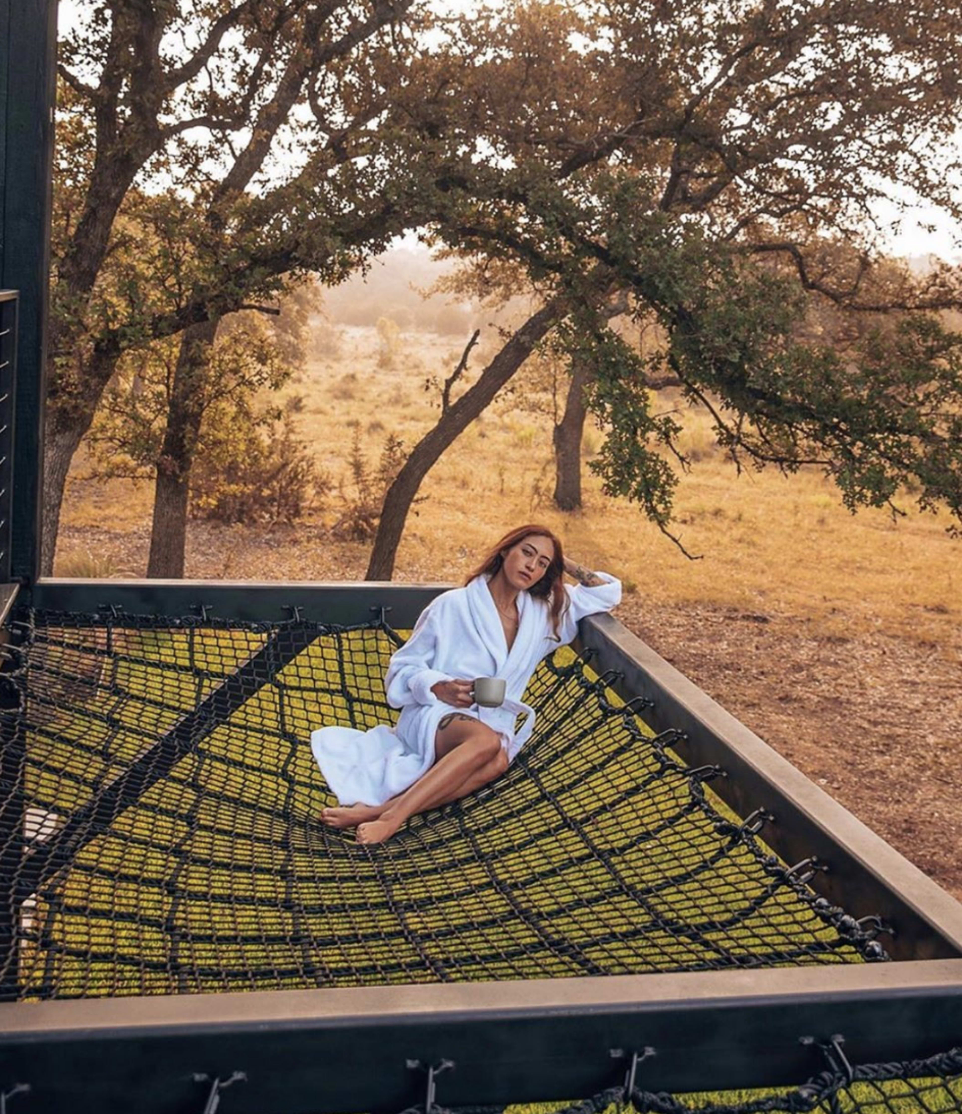 A person in a white robe is sitting on a large net suspended between trees in a natural setting, holding a drink.
