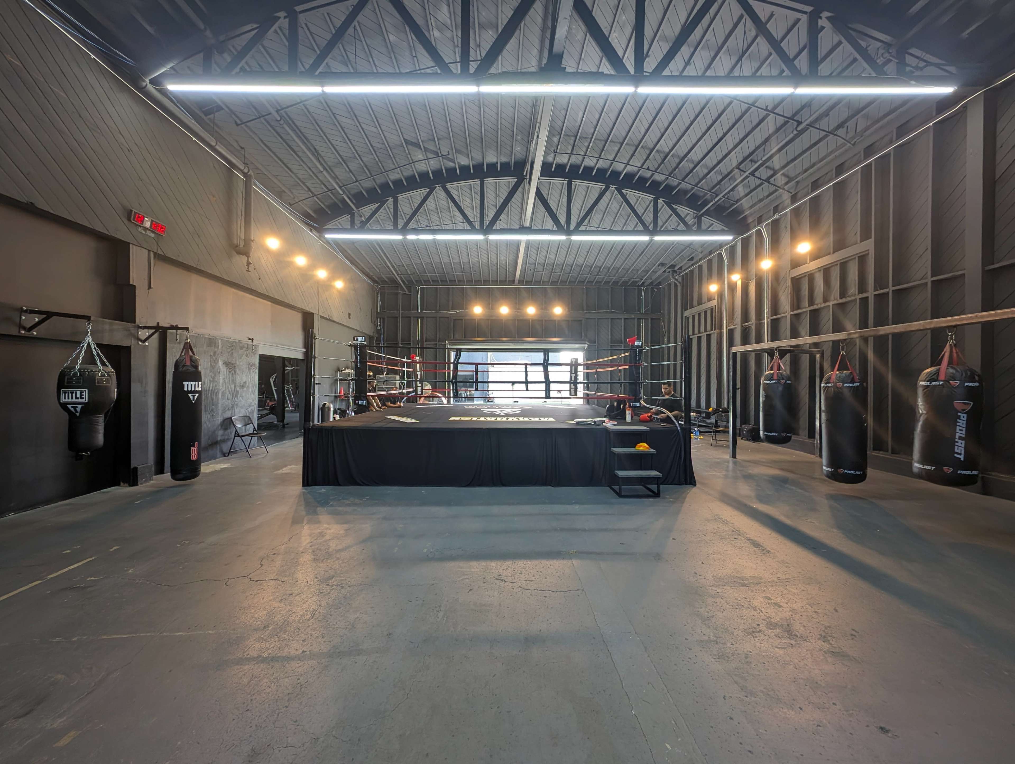 A spacious boxing gym featuring a central ring, heavy punching bags hanging from the walls, and bright overhead lighting.