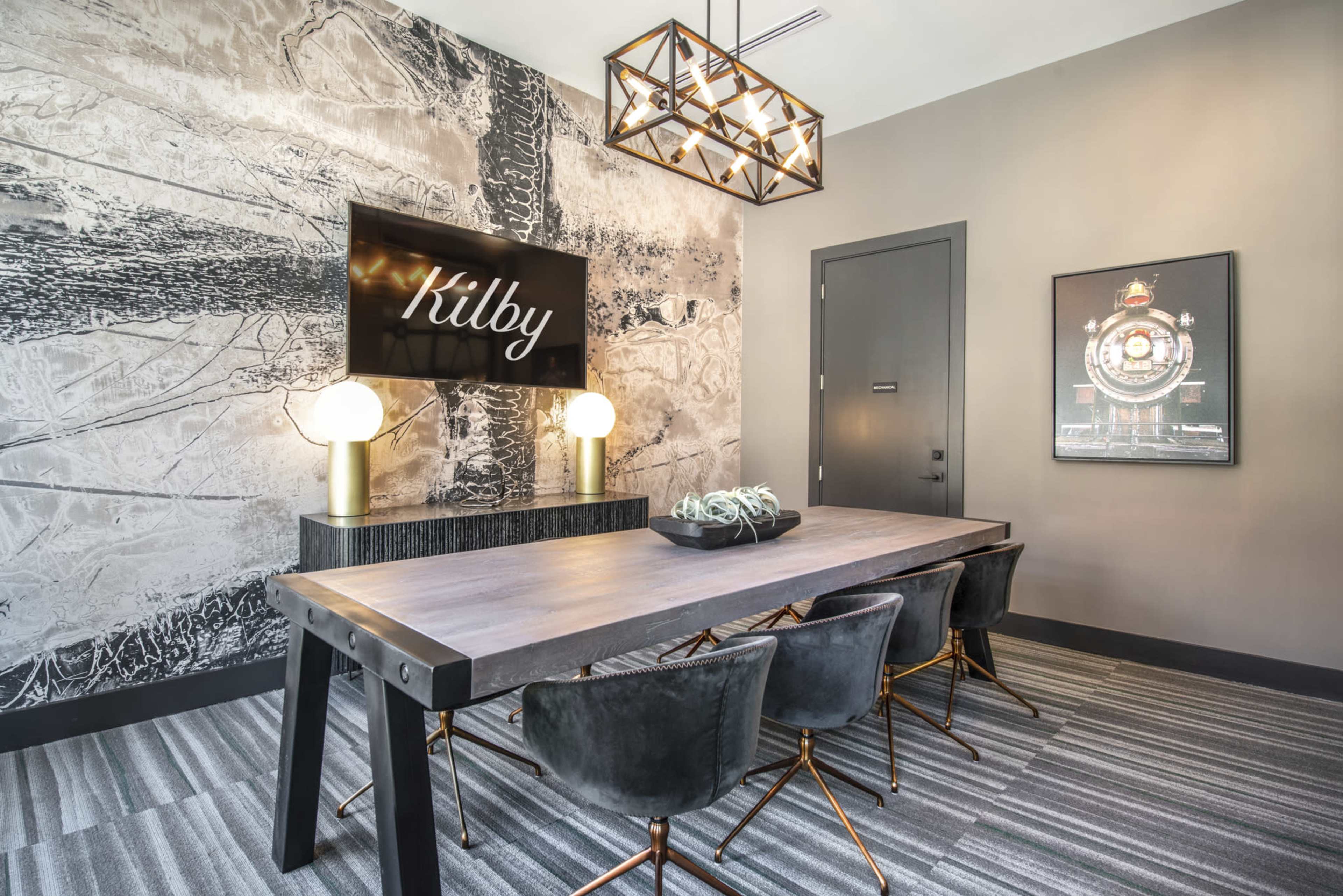 A modern conference room features a large wooden table surrounded by dark chairs, with a decorative wall and a screen displaying the name "Kilby."