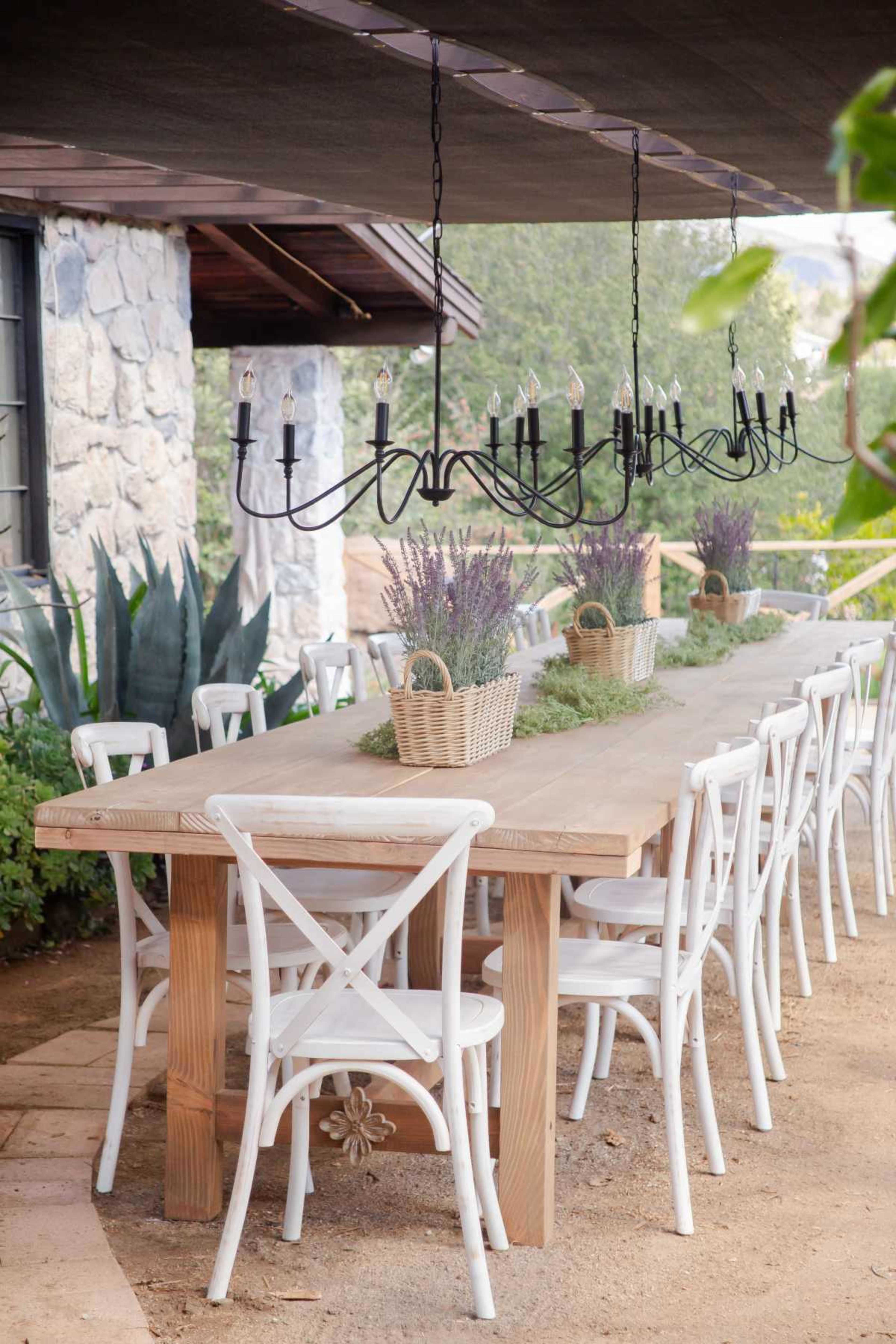 A long wooden dining table is set outdoors under a covered area, surrounded by white cross-back chairs and adorned with centerpieces of lavender in woven baskets.