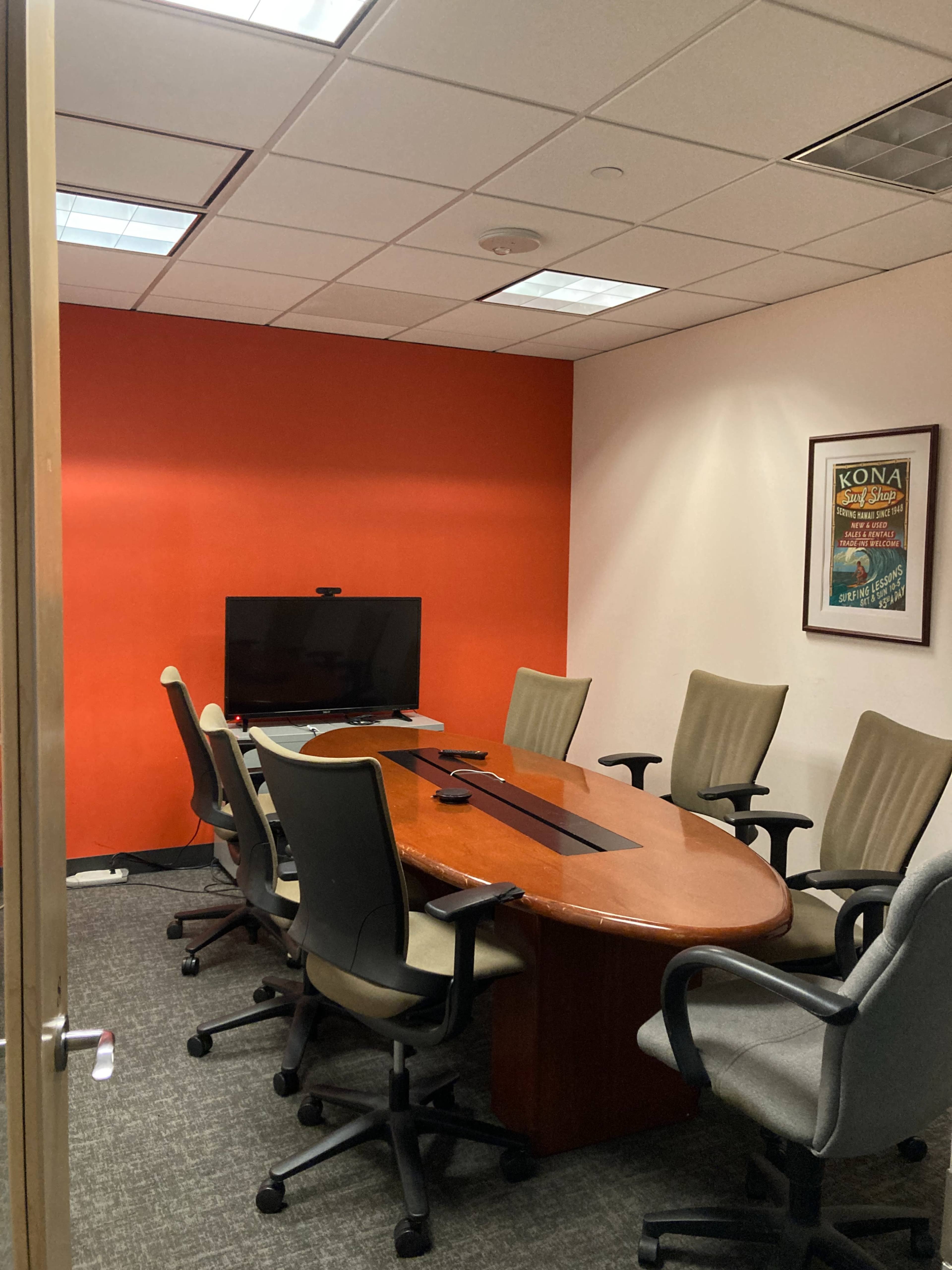 A small conference room features a large wooden table surrounded by six office chairs and a wall-mounted TV on an orange wall.