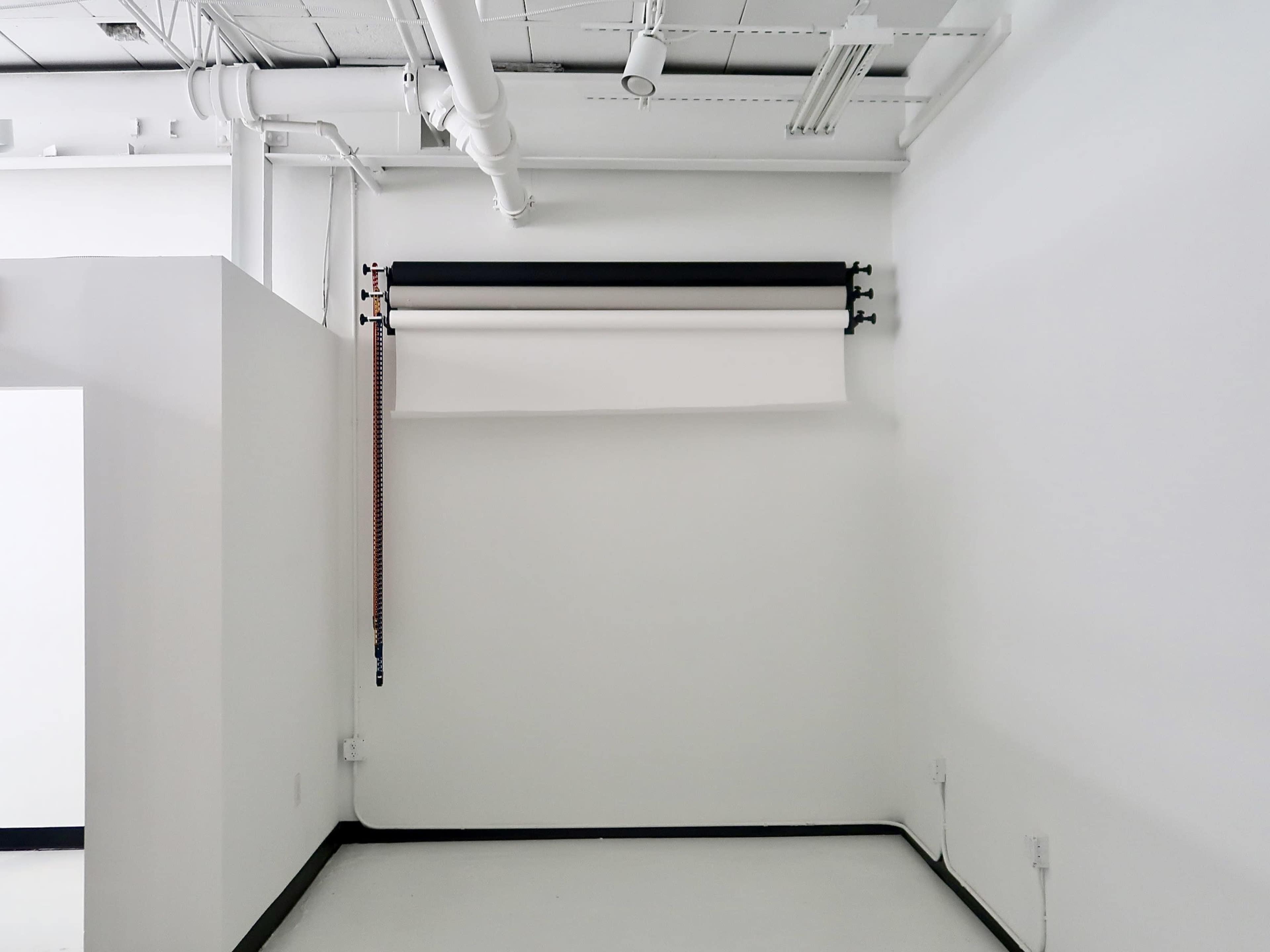 The image shows a bare corner of a room with a white wall, featuring a roll-up backdrop system mounted near the ceiling.