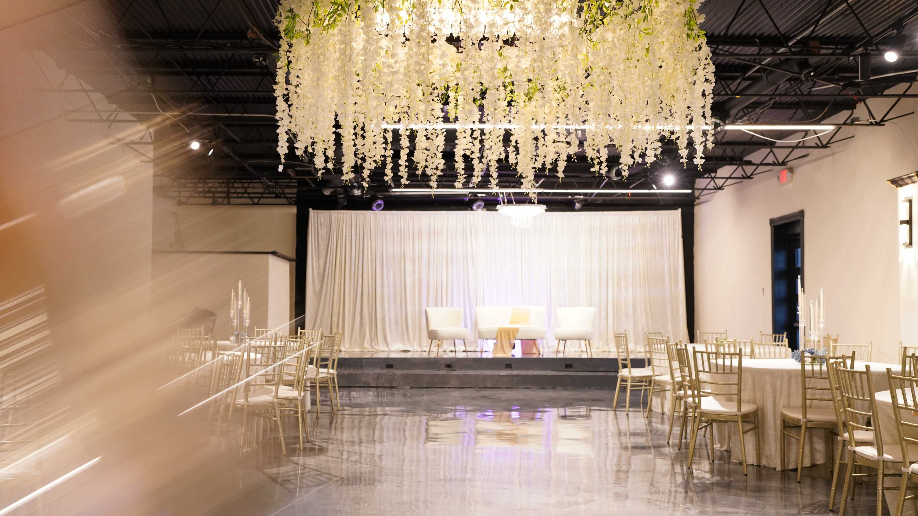 The image shows an elegantly arranged banquet hall with gold chairs, round tables, and a decorative ceiling featuring hanging floral elements.