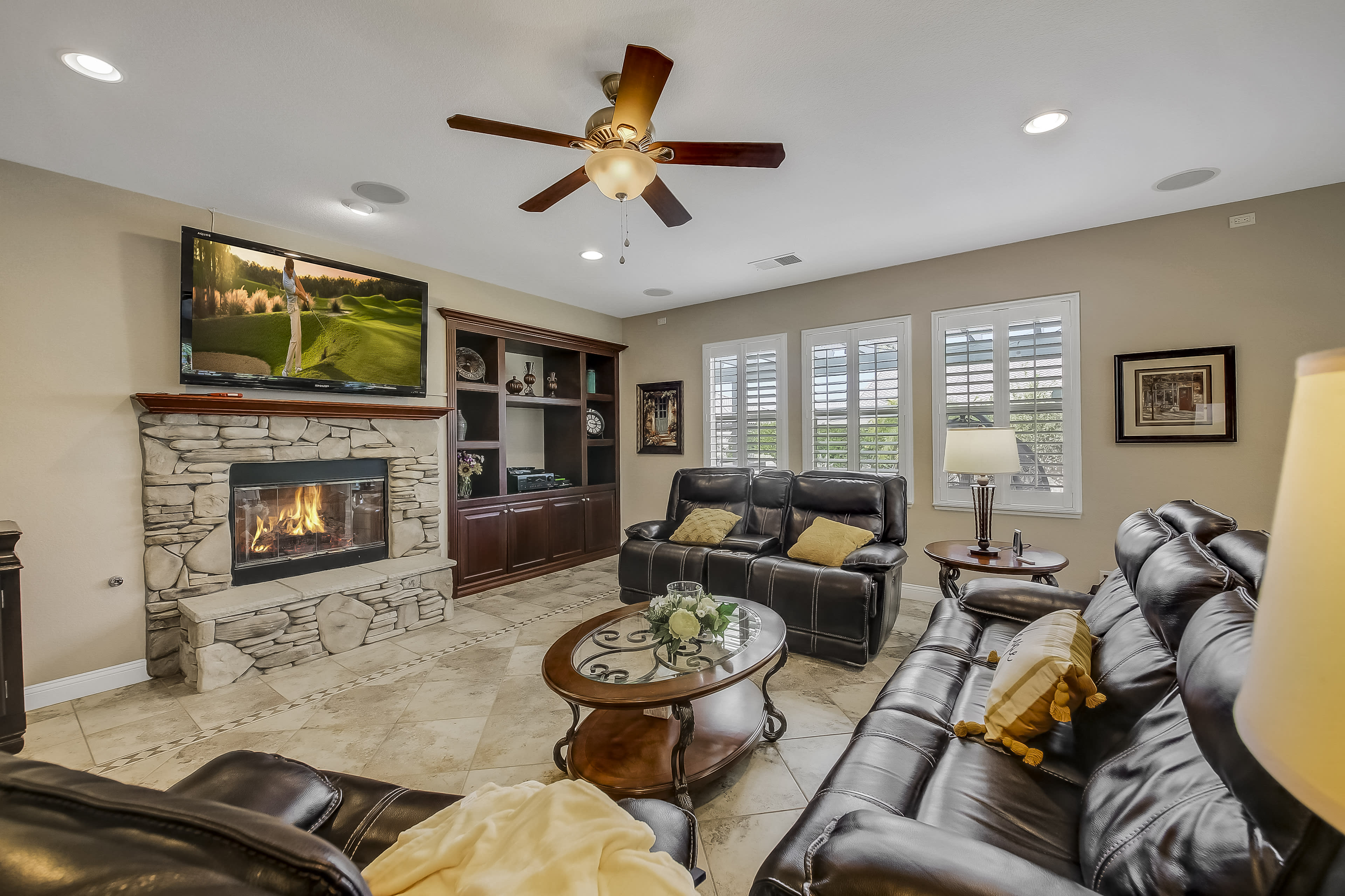 The image shows a living room with a stone fireplace, a large television mounted above it, and leather sofas arranged around a circular coffee table.