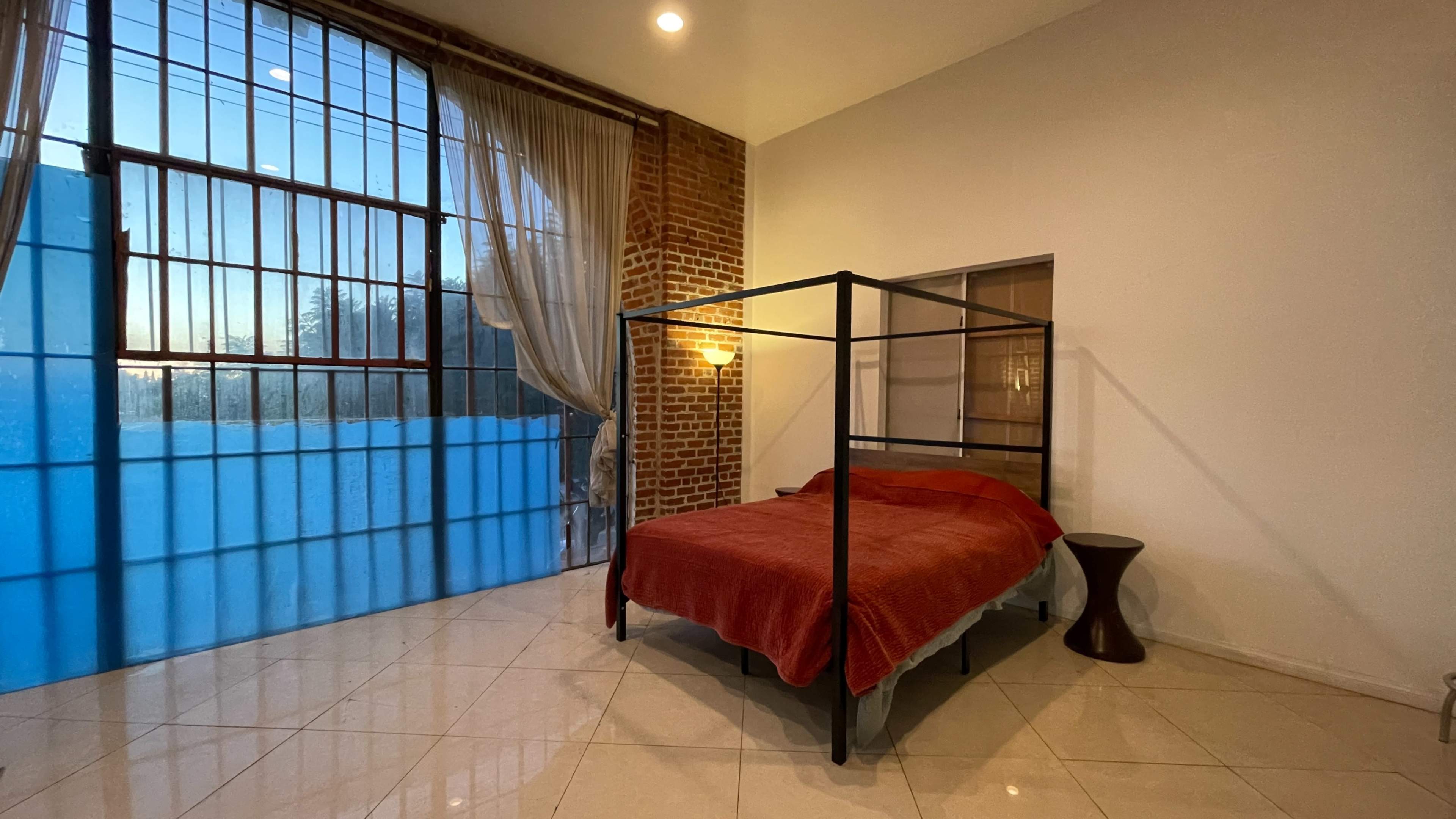 A bed with a red cover is positioned in a room featuring large windows with blue curtains and exposed brick walls.
