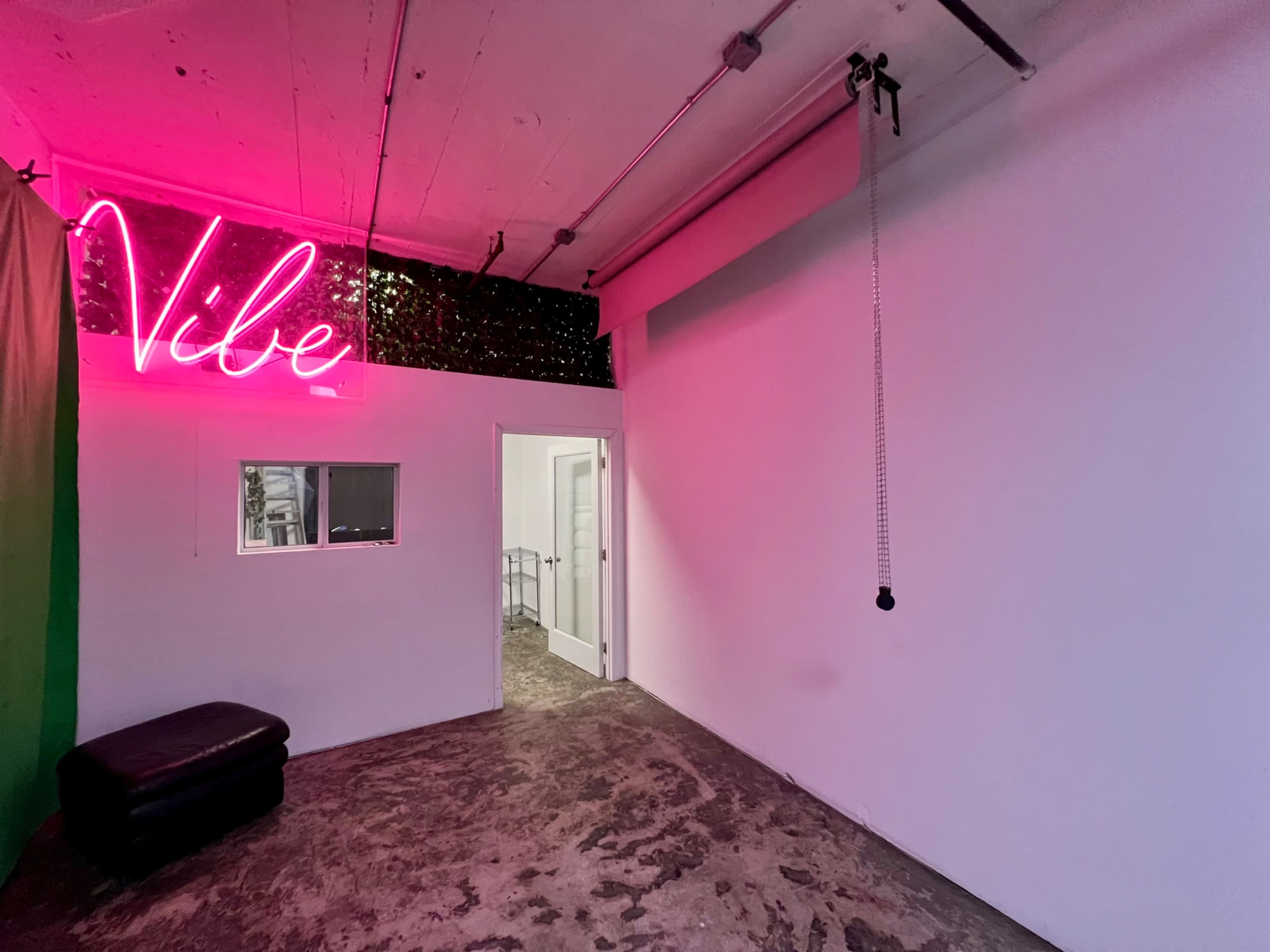 A spacious room features a neon "Vibe" sign, a small window, and an open doorway leading to another area.