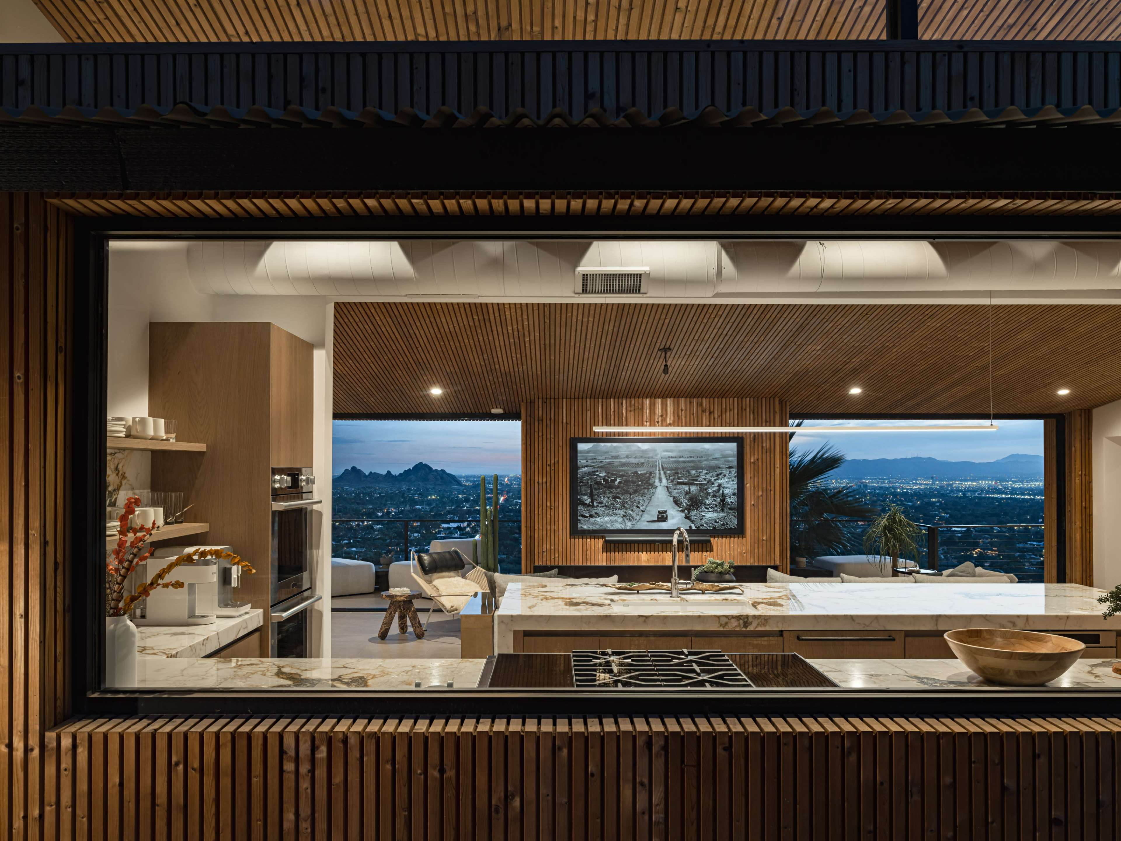 A modern kitchen with wooden cabinetry and a large window revealing a view of a landscape with mountains and a city skyline in the distance.