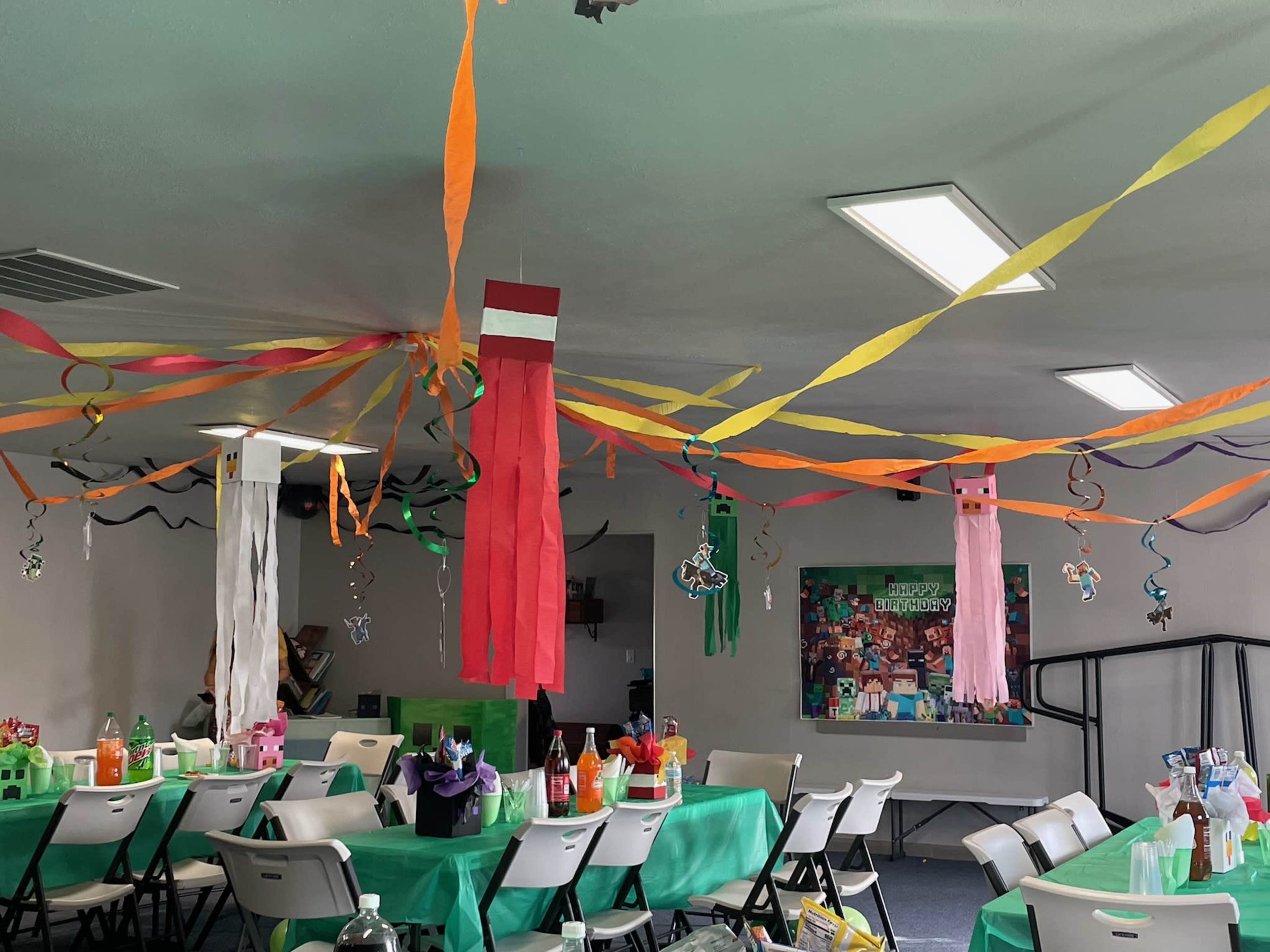 The room is decorated for a party with colorful streamers hanging from the ceiling and multiple tables set with drinks and party supplies.