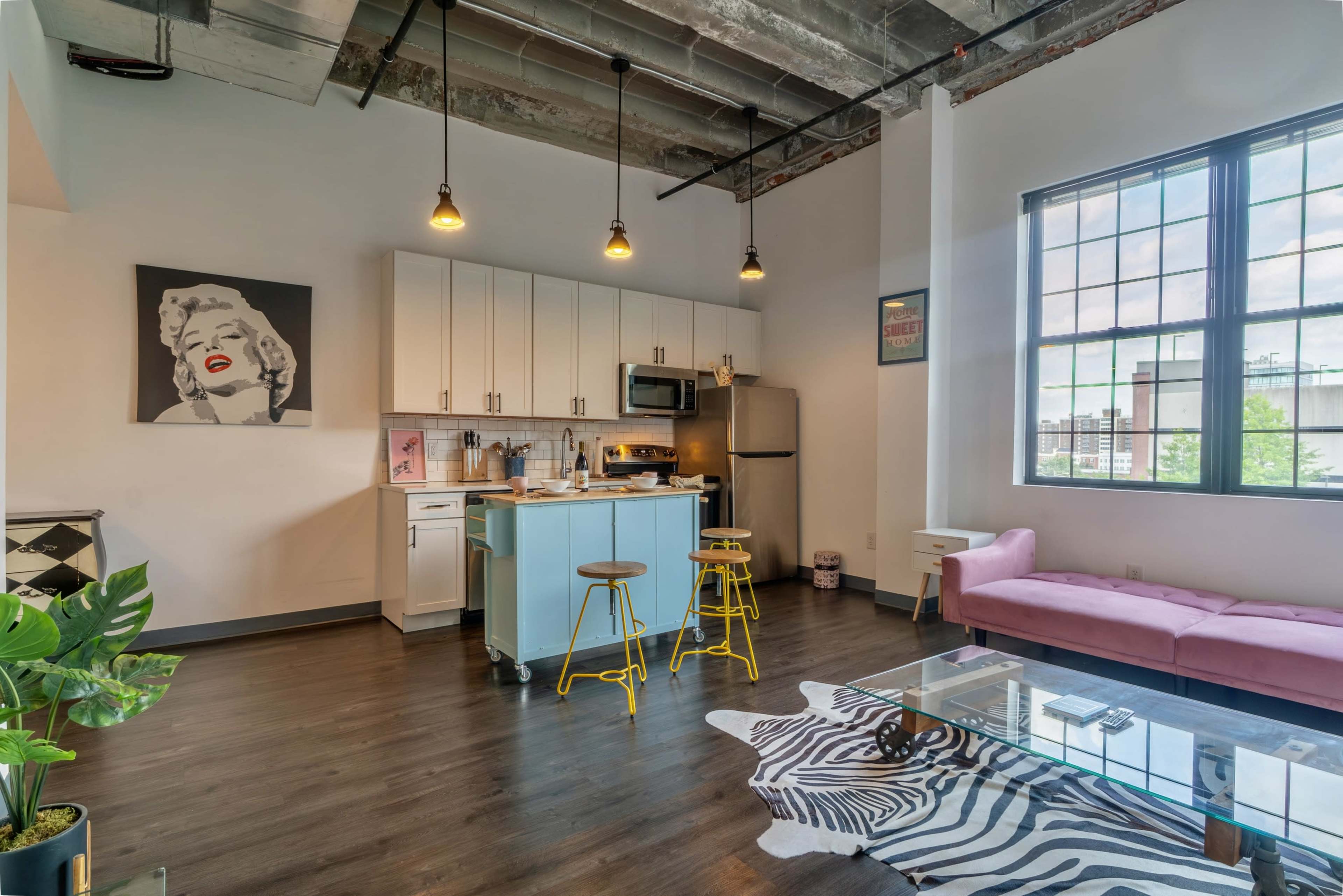 The image shows a modern apartment interior featuring a kitchen with a blue island and two stools, a living area with a window, and decorative elements including a zebra-print rug and a portrait of a woman on the wall.